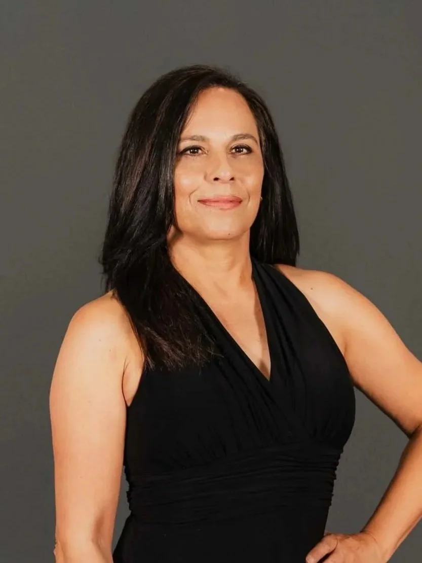 A woman with medium-length dark hair wearing a sleeveless black dress, standing against a plain gray background, smiling with one hand on her hip.