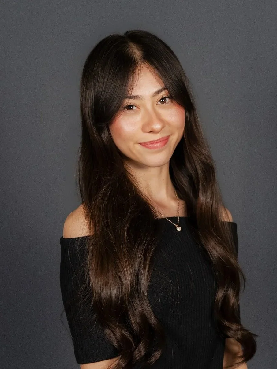 A woman with long dark brown hair, wearing a black off-the-shoulder top and a necklace, smiling at the camera against a gray background.