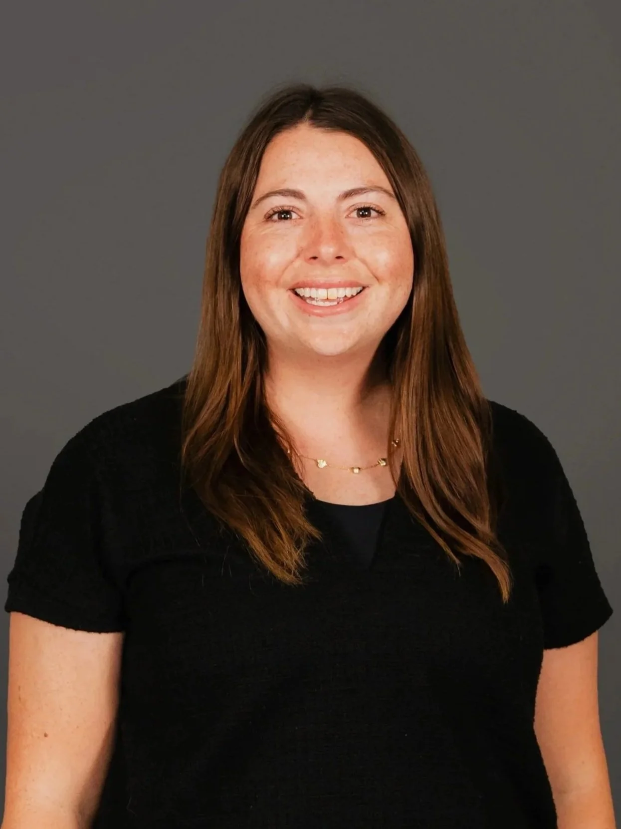 A woman with long brown hair and a big smile, wearing a black top and a gold necklace, against a gray background.