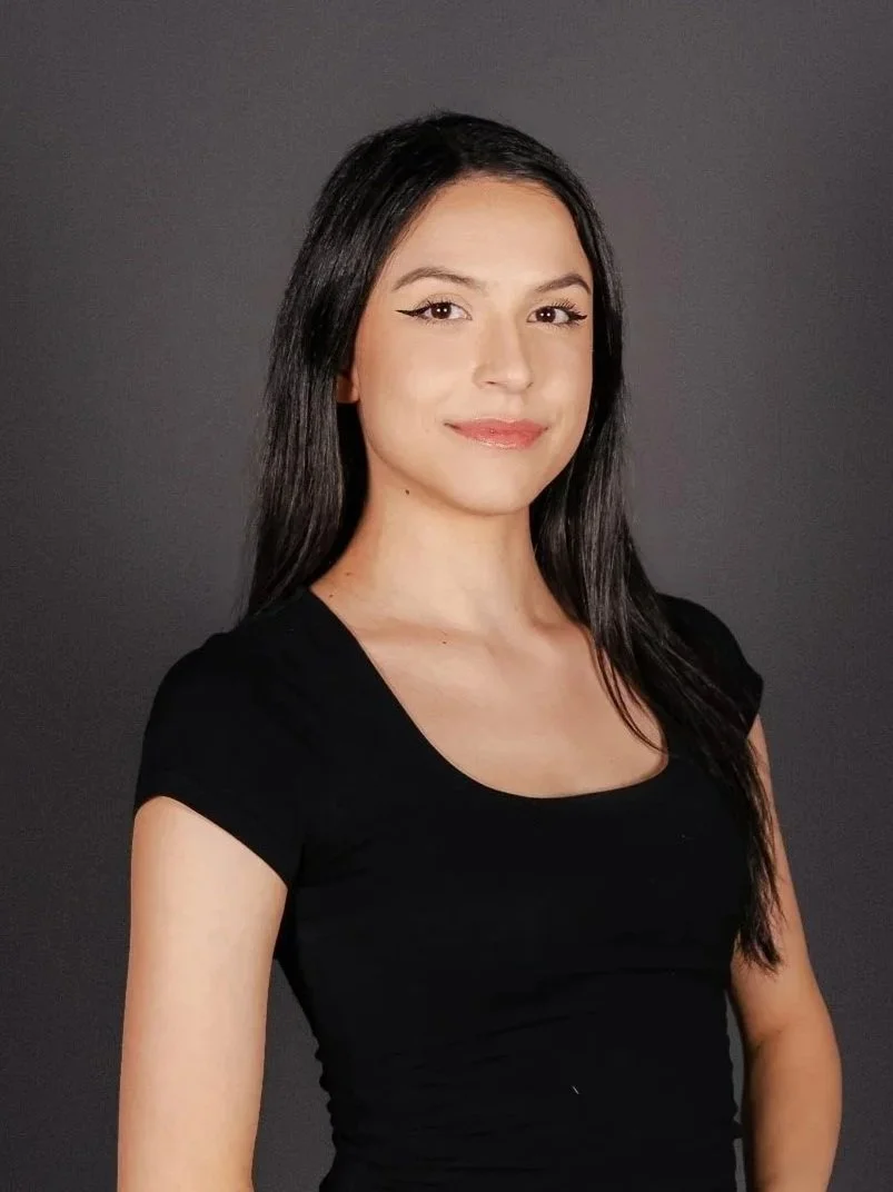 A young woman with long black hair, wearing a black top, smiling softly against a dark gray background.