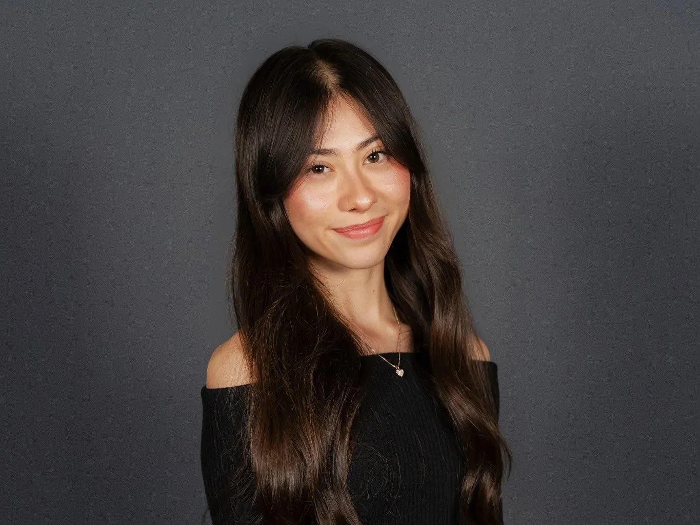 A woman with long dark brown hair, wearing a black off-the-shoulder top and a necklace, smiling at the camera against a gray background.