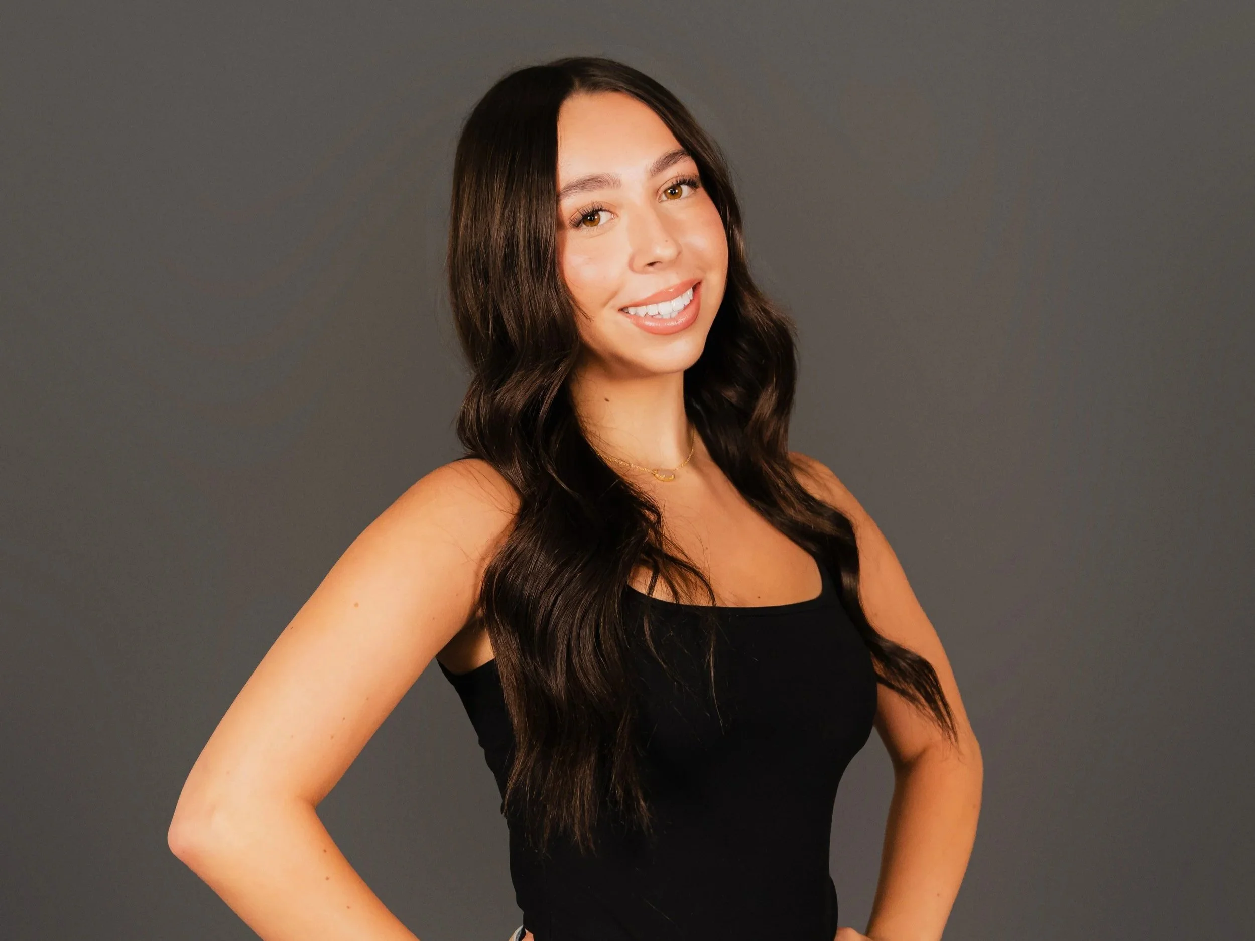A young woman with long dark hair, wearing a black tank top, smiling, standing against a plain gray background.