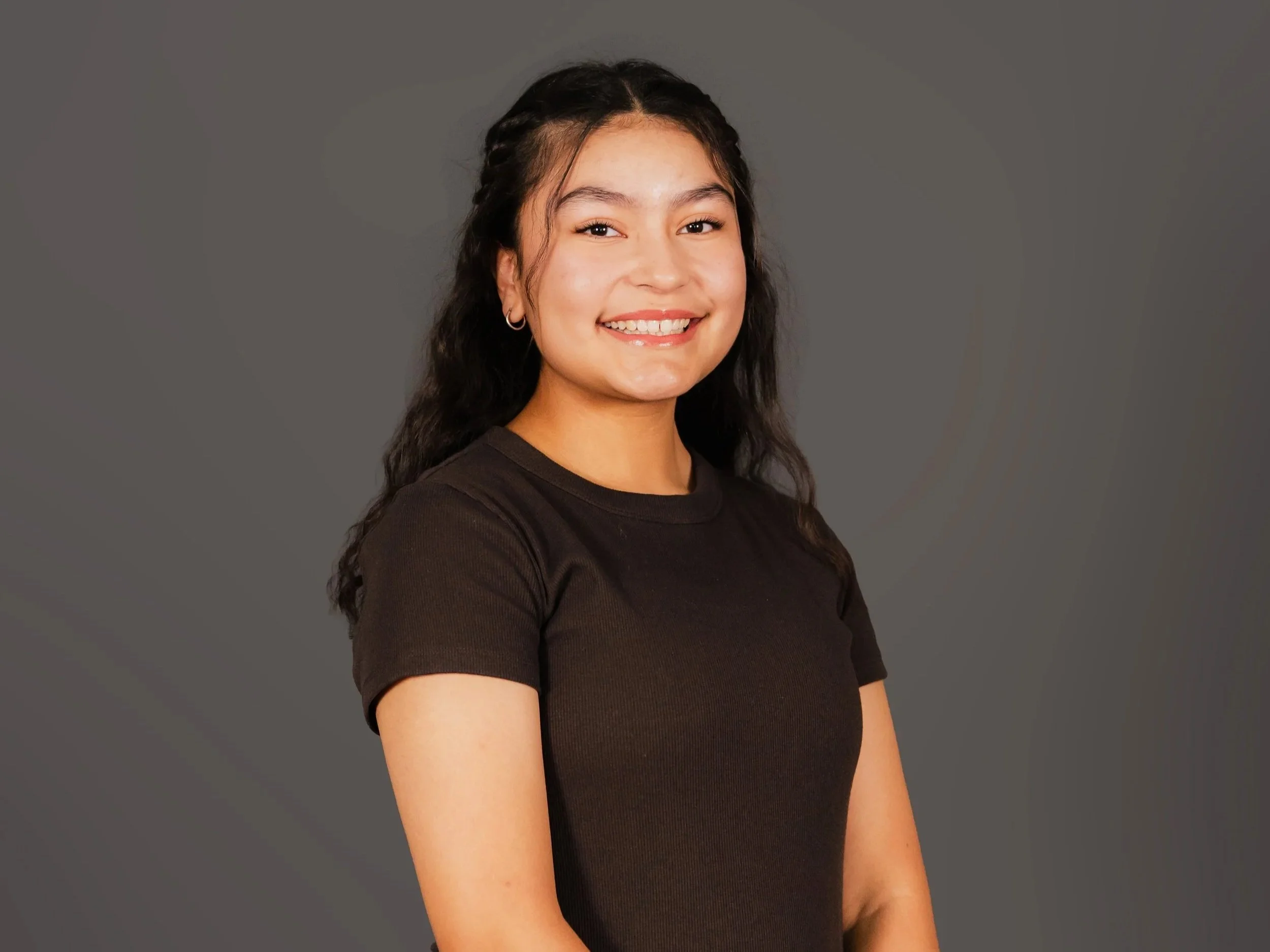 A young woman with long dark hair styled in braids, smiling and wearing a black short-sleeve shirt, standing against a plain gray background.