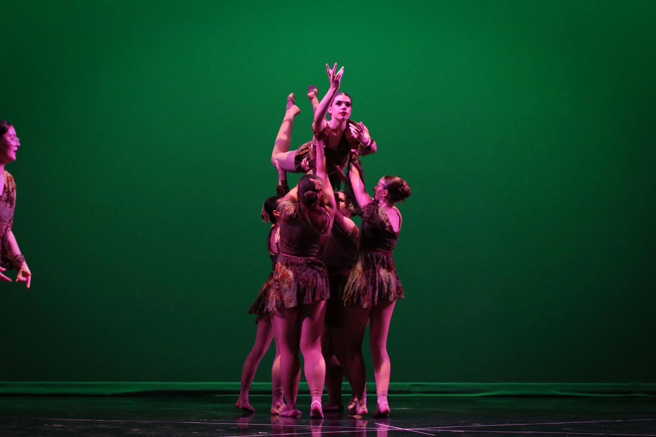 A group of ballet dancers perform a lift on stage with a green backdrop. One dancer is being held up horizontally by her fellow dancers, with her arms extended and focused expression.