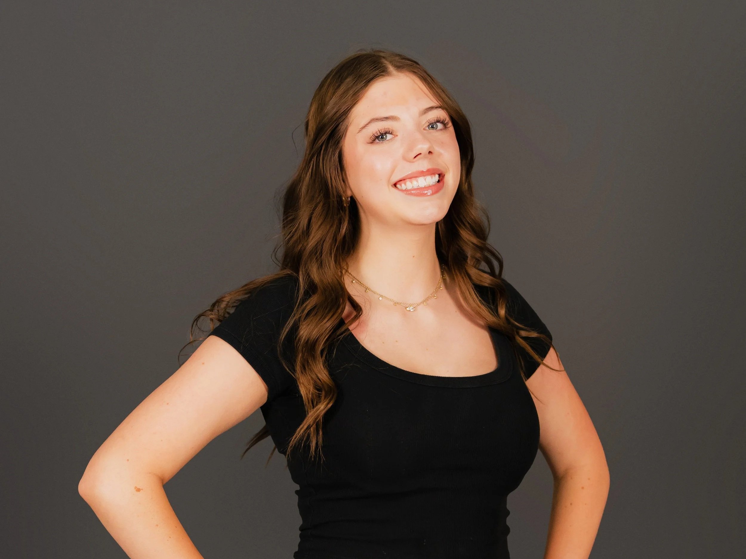A young woman with long wavy brown hair and blue eyes smiling, wearing a black top and layered gold necklaces, standing against a plain gray background.