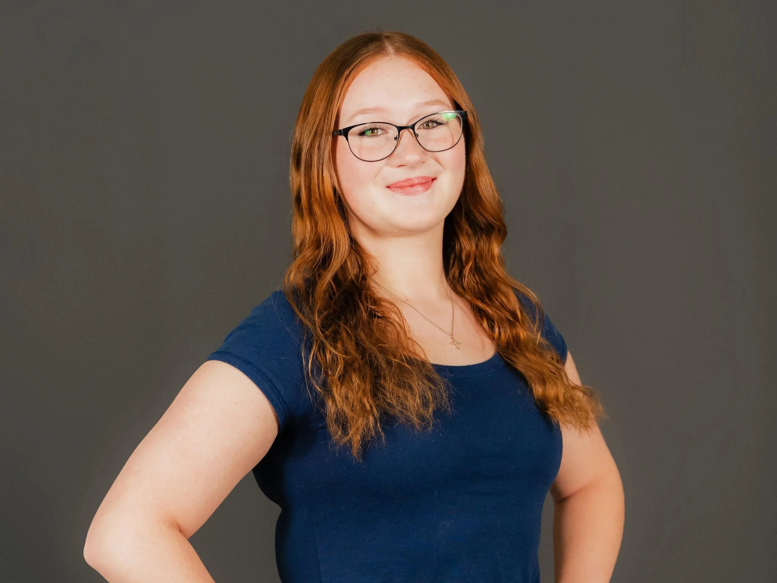 A young woman with red hair, glasses, and a blue shirt, smiling against a gray background.