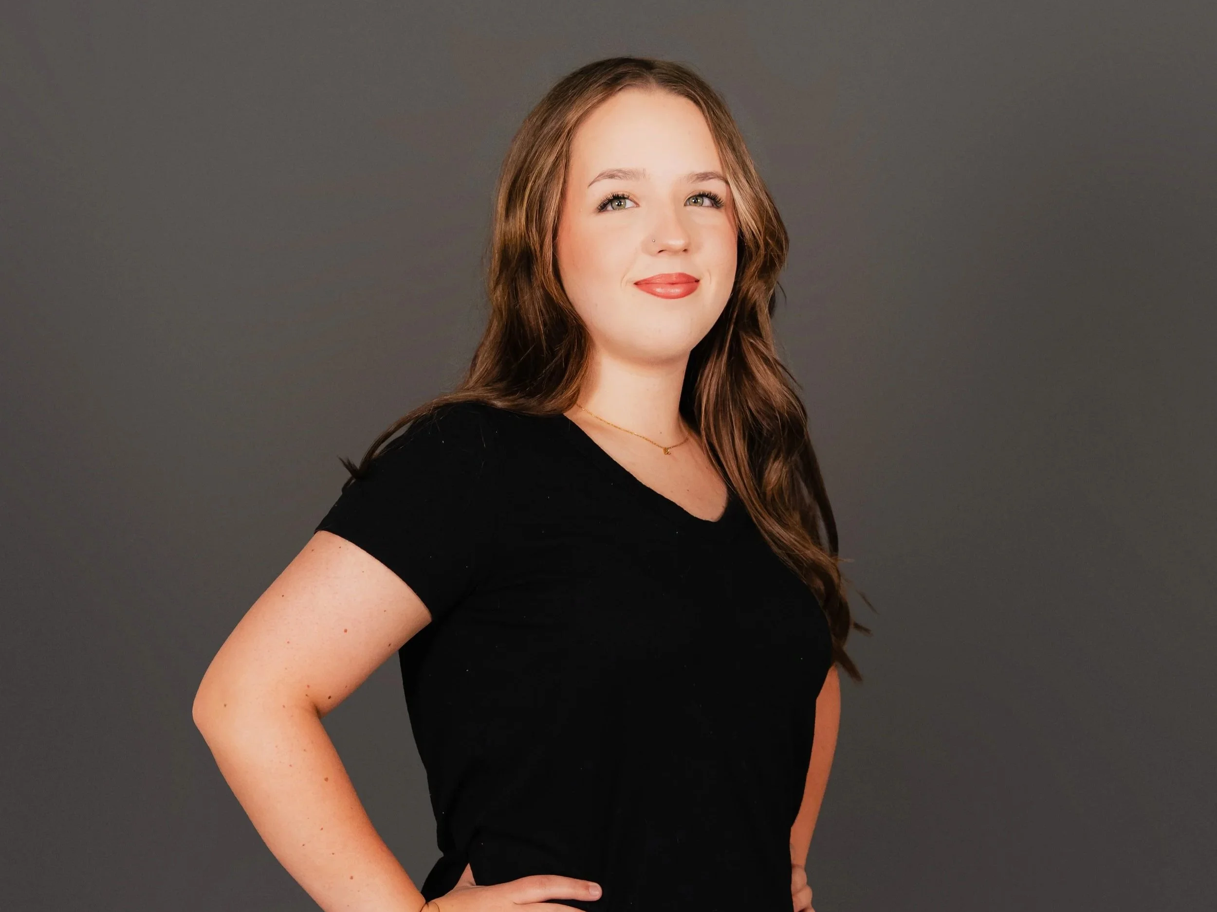 A woman with long brown hair, wearing a black top, posing against a plain grey background.