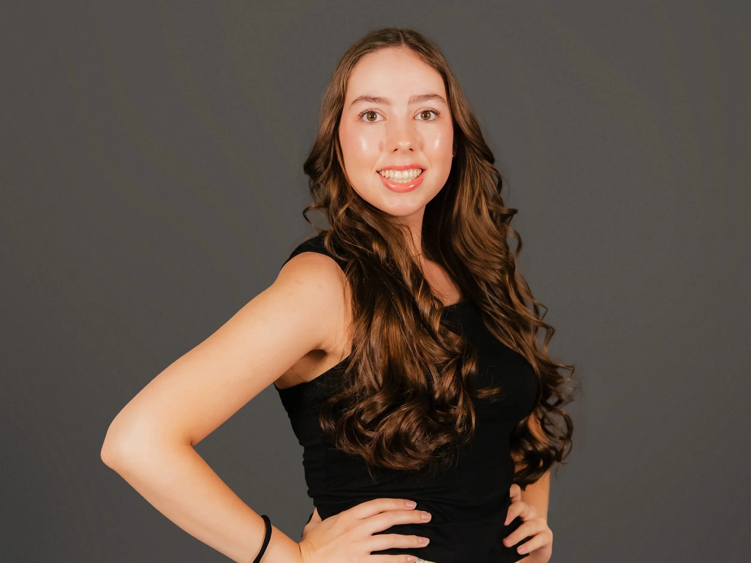 A young woman with long, curly brown hair, smiling and looking at the camera, wearing a black sleeveless top and standing against a gray background.