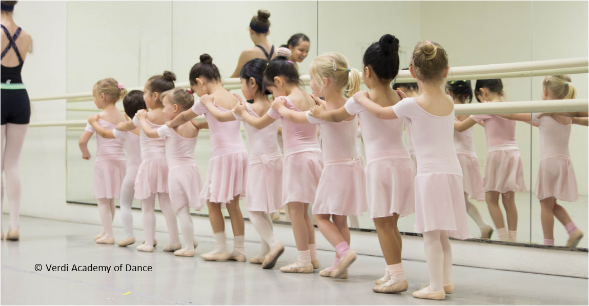 Young ballet dancers in pink leotards and skirts holding onto each other's shoulders in a line.
