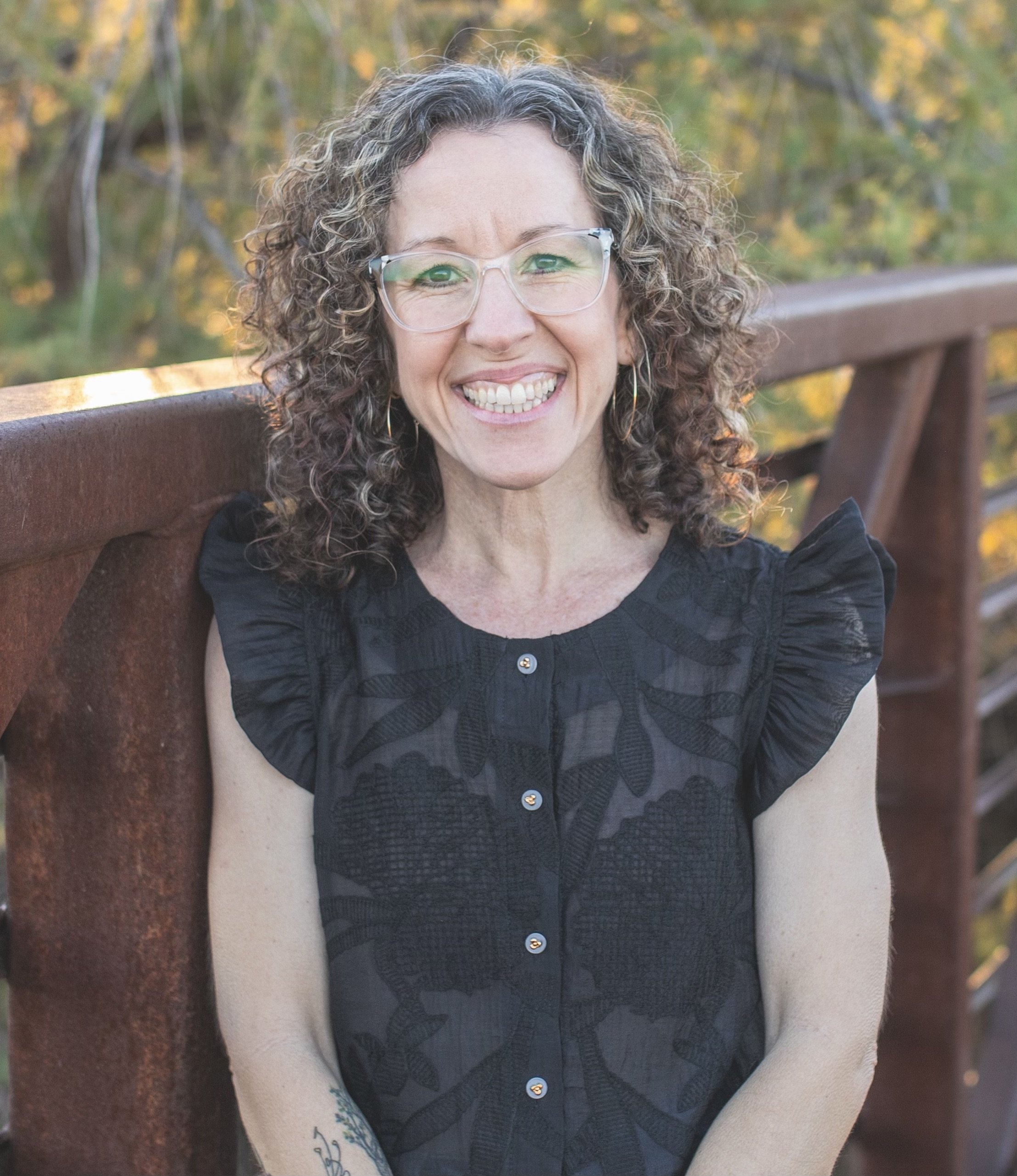 A woman with curly hair smiling and leaning against a wall in a sleeveless patterned top.