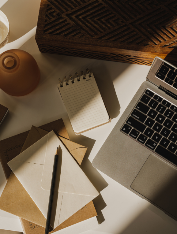 Close-up of a workspace with a laptop, a small notepad, envelopes, a pen, and a decorative wooden box on a white table, with sunlight casting shadows.