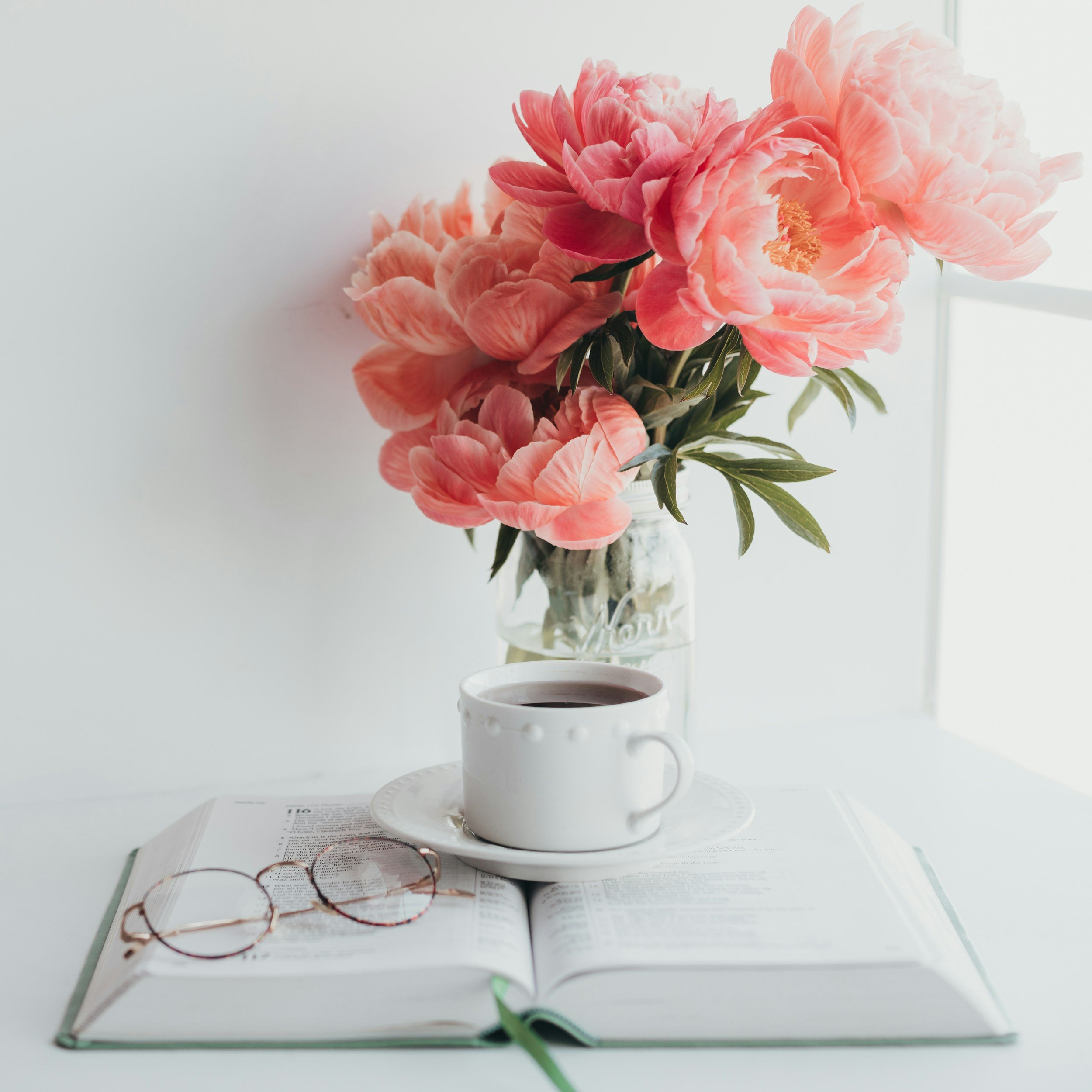 A vase with pink peony flowers, a white coffee cup of coffee, a pair of reading glasses, and an open book with text and a green bookmark on a white surface.