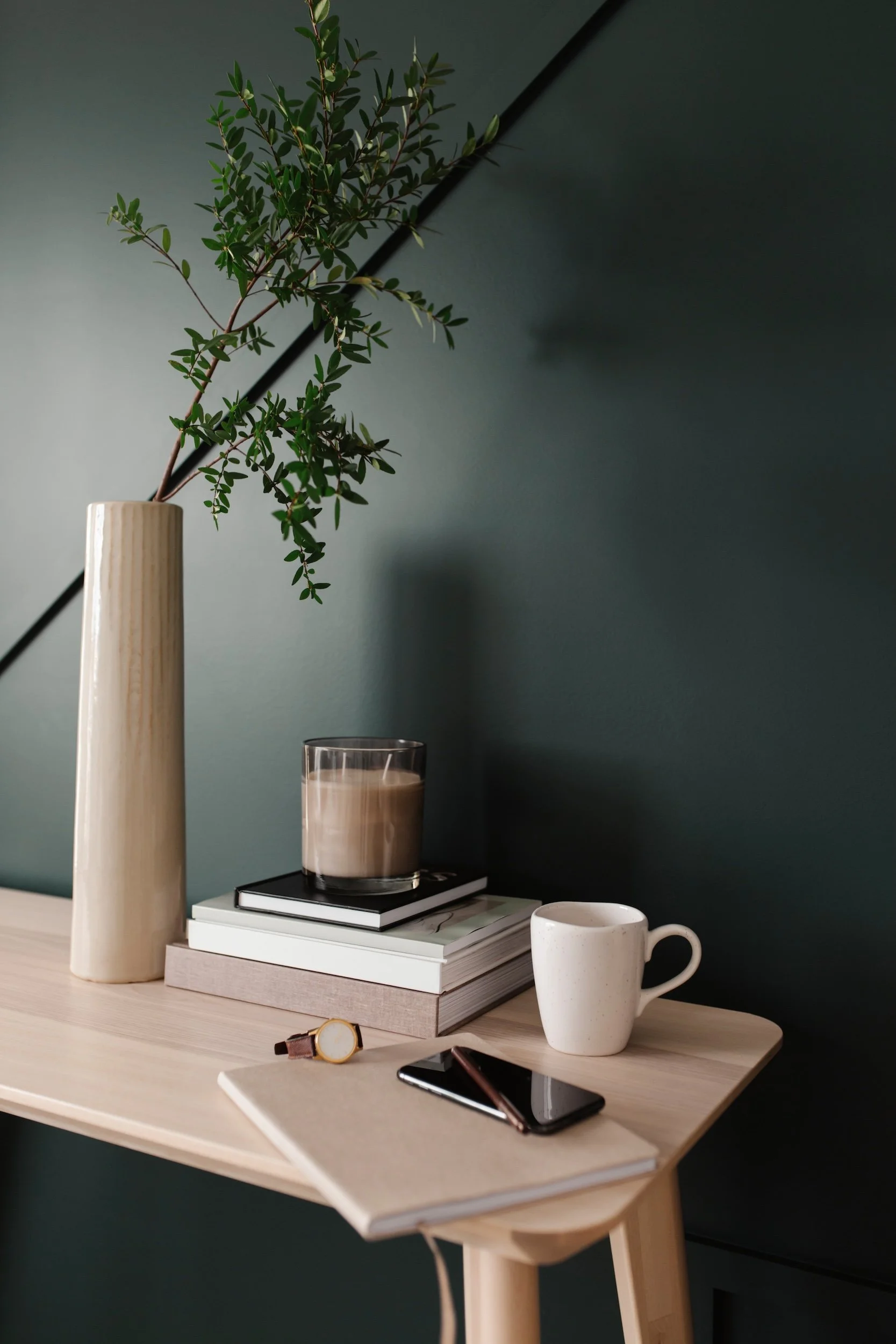 A desk with a tall beige vase holding green leafy branches, stacked books, a glass candle with beige wax, a white speckled coffee mug, a black smartphone, a gold watch, and two closed notebooks, set against a dark teal wall.