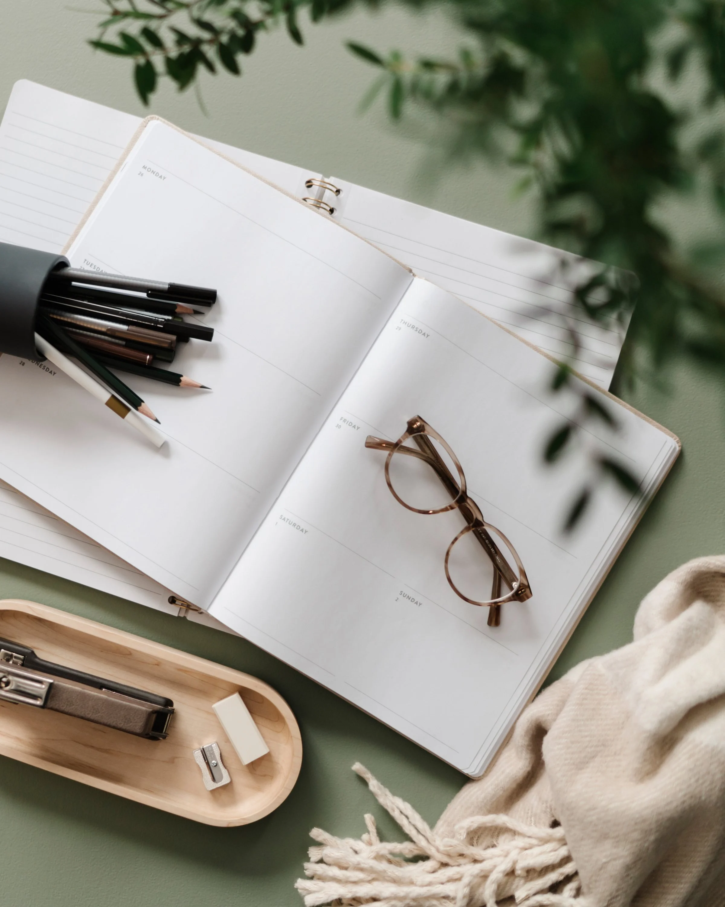 Open planner with weekly pages, black pens, a pair of glasses, and a wooden box containing a stapler and small stationery items on a green surface.