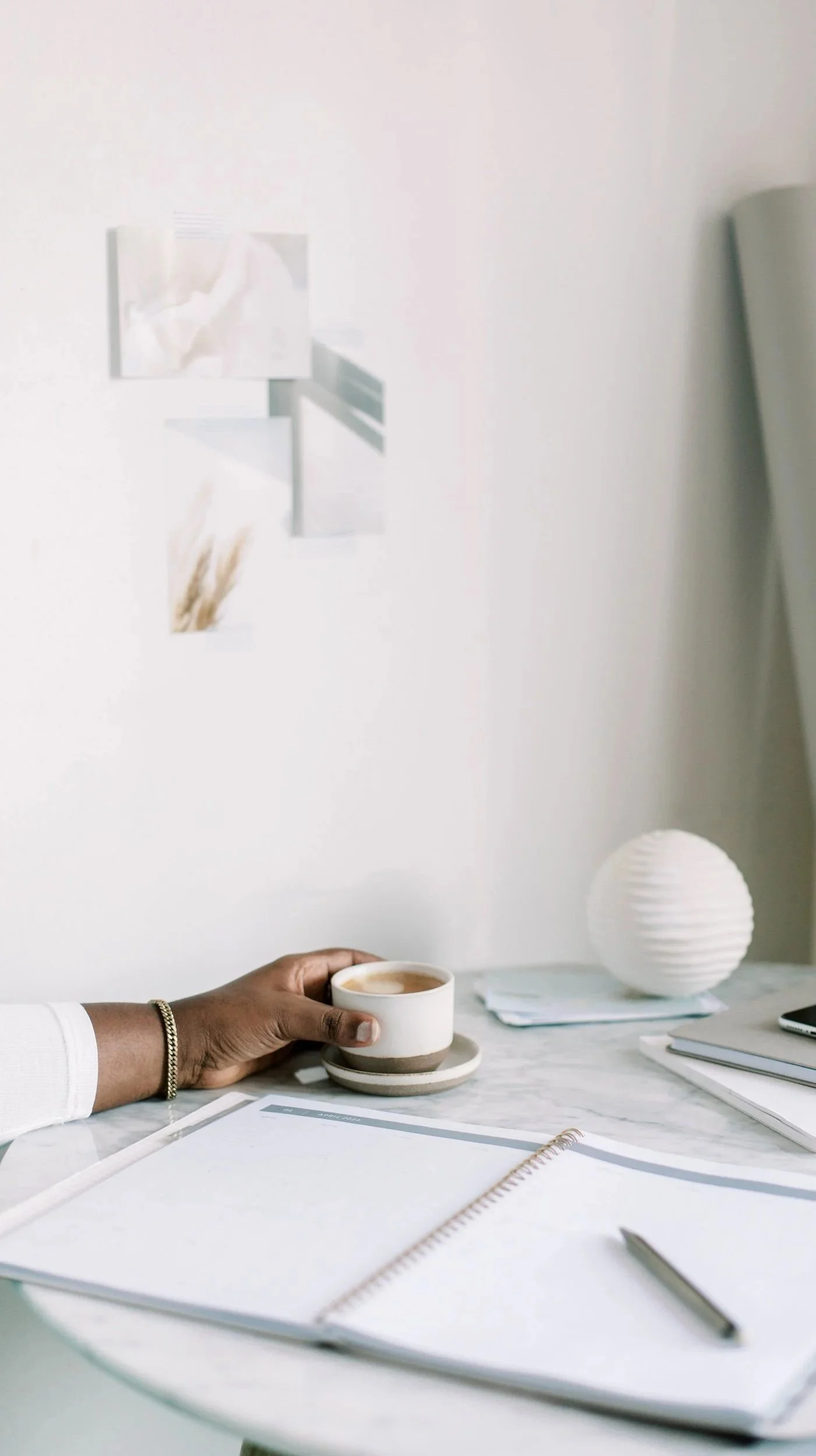 A person's hand holding a small coffee cup with a saucer on a white marble table. On the table are notebooks, a pen, and a paper. The background has a white wall with three small abstract art pieces and a white round paper lamp.