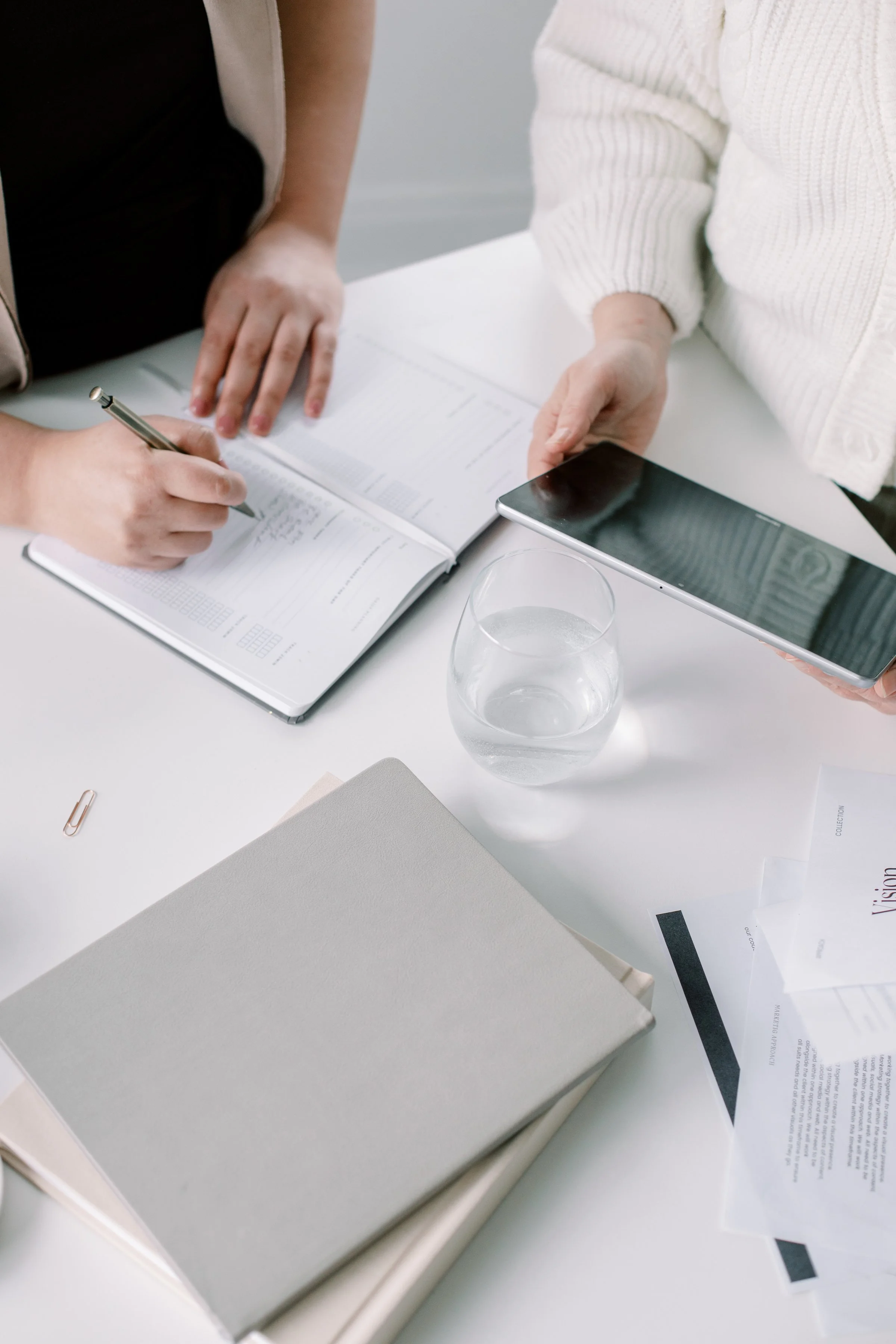 People working at a table with documents, a glass of water, a smartphone, and a closed white book.
