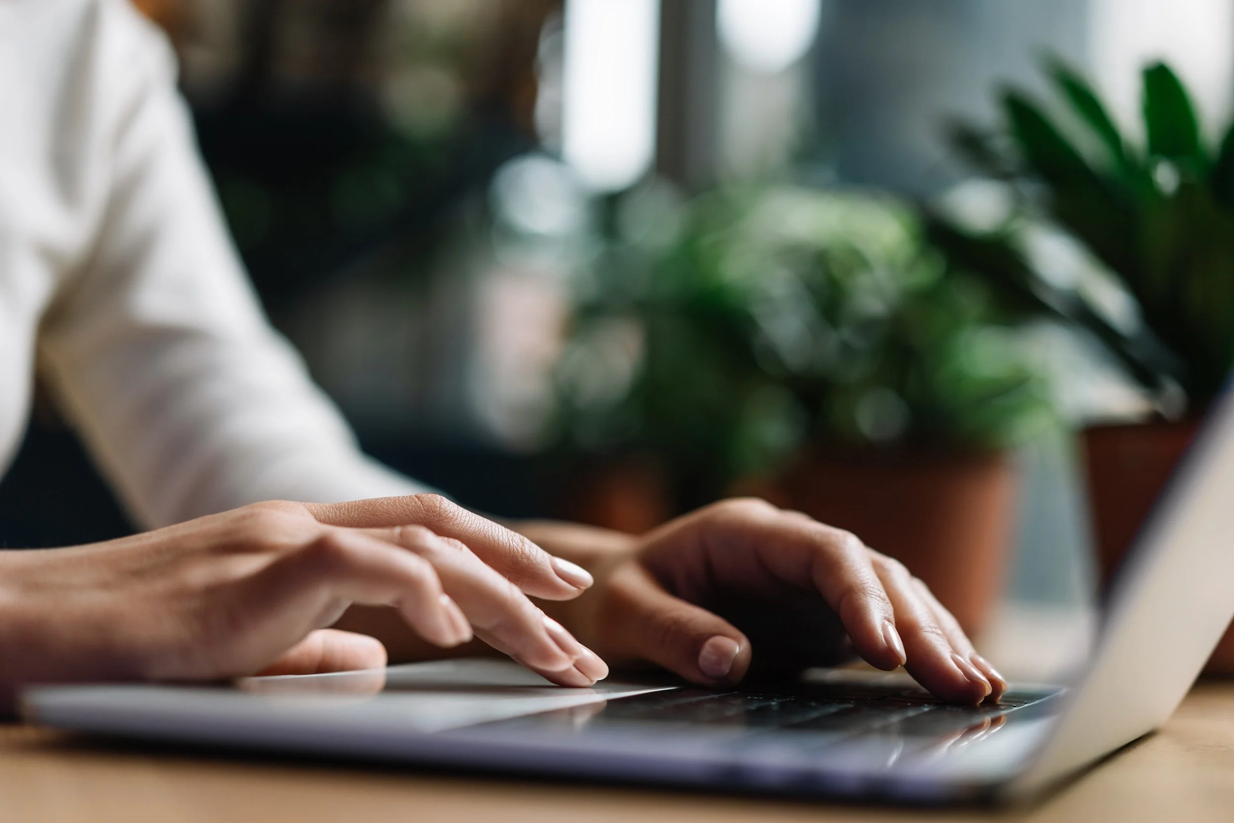Close-up of hands typing on a laptop keyboard with a blurred background of indoor plants.