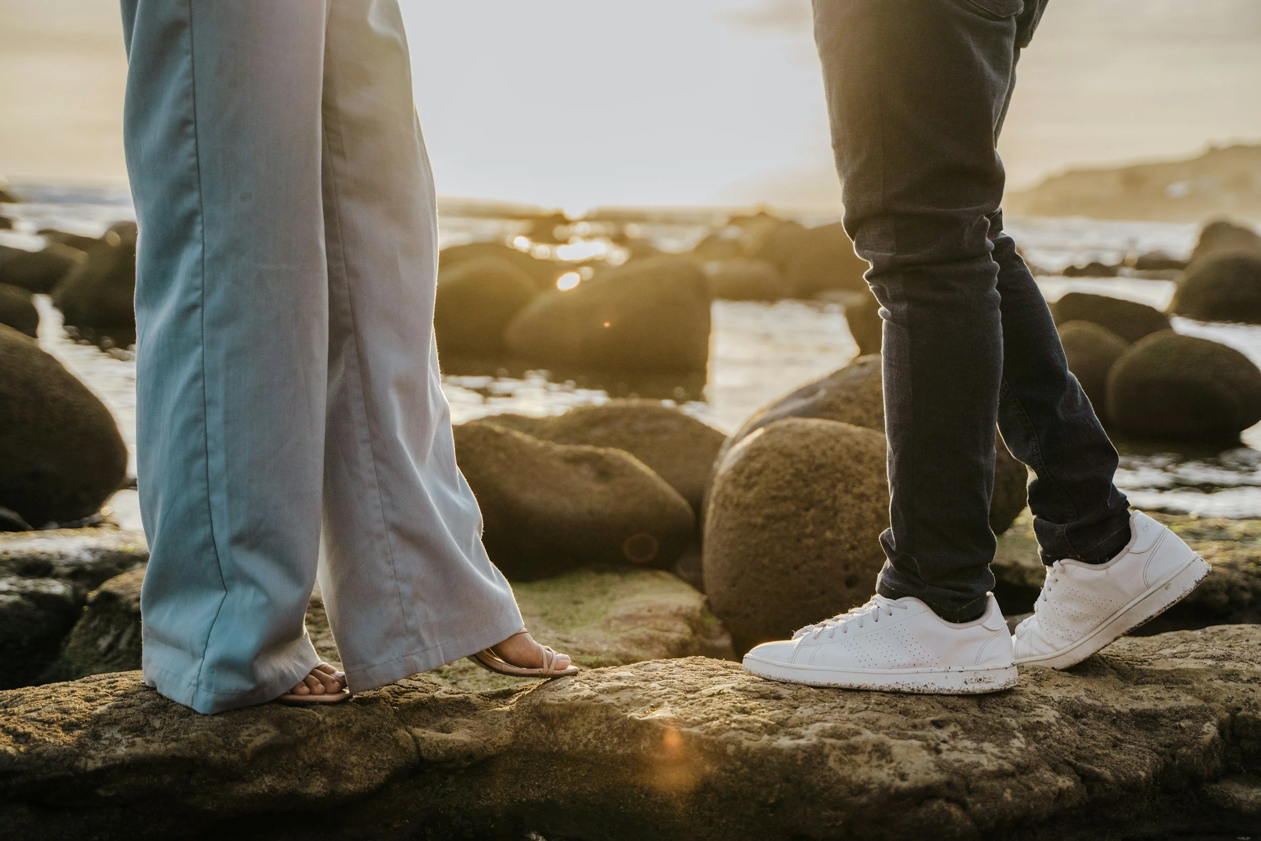 Close-up of two people standing on rocks by the water during sunset, showing their legs and shoes. One is wearing beige pants and sandals, the other is wearing dark jeans and white sneakers.