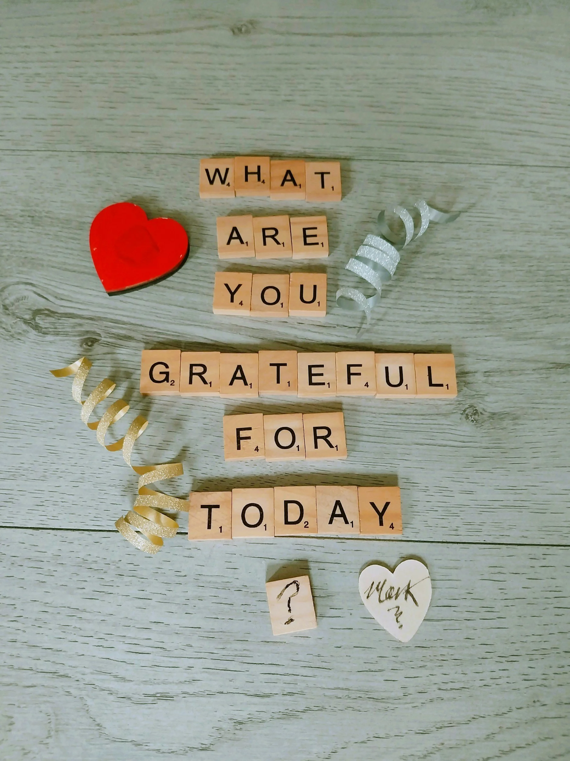 A wooden surface with alphabet tiles spelling out "What are you grateful for today," a red heart, a small question mark, and a heart-shaped note with "Work?" written on it, along with decorative curly ribbons.
