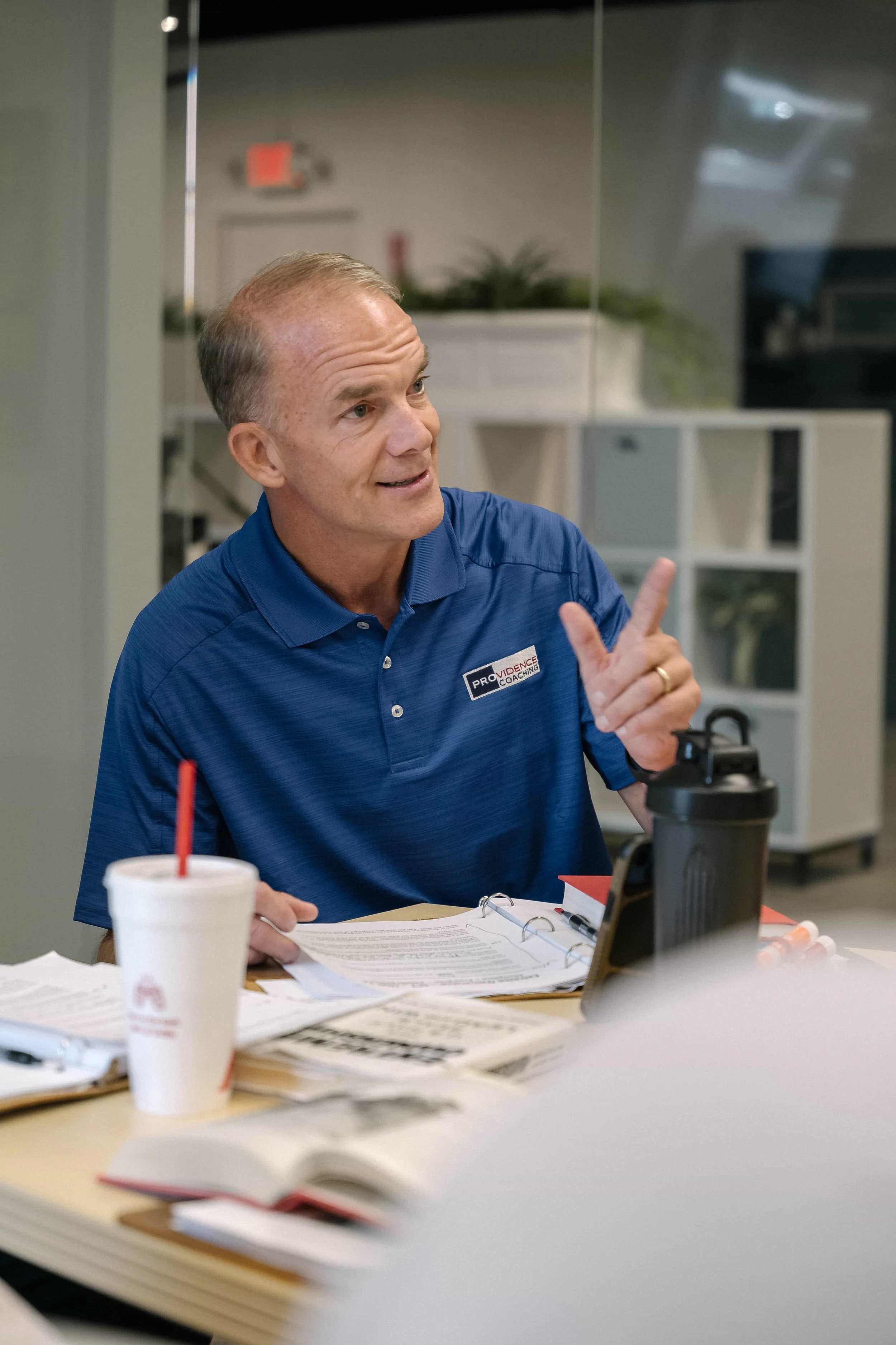 A man in a blue polo shirt is gesturing with his right hand during a meeting or discussion. He is seated at a table cluttered with papers, a notebook, and drinks. The background shows office furniture and plants, with a glass wall or partition.