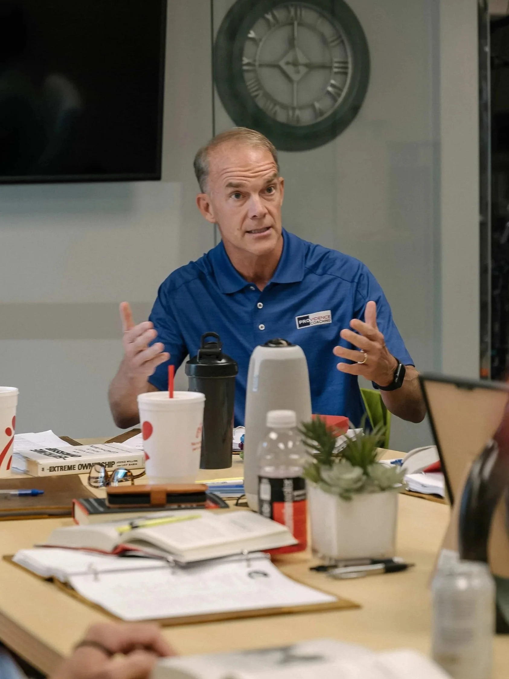 A man in a blue shirt gesturing with his hands, sitting at a table with notebooks, papers, cups, and pens, in a room with a large clock on the wall behind him.