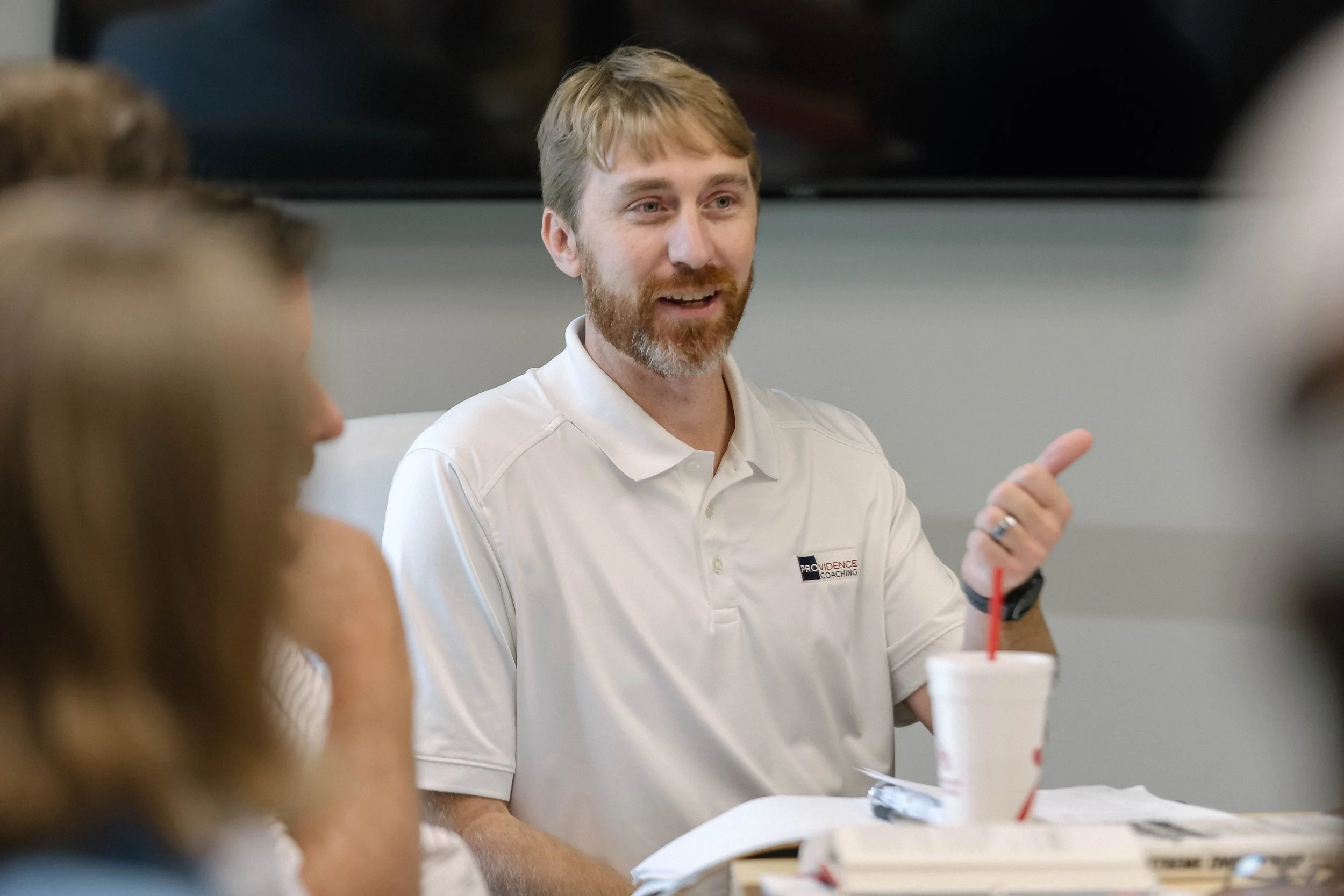 A man with a beard and light brown hair, wearing a white polo shirt with a Providence Coaching logo, is speaking or explaining something in a meeting or discussion. There are blurred people in the foreground, and a drink with a red straw is on the table.