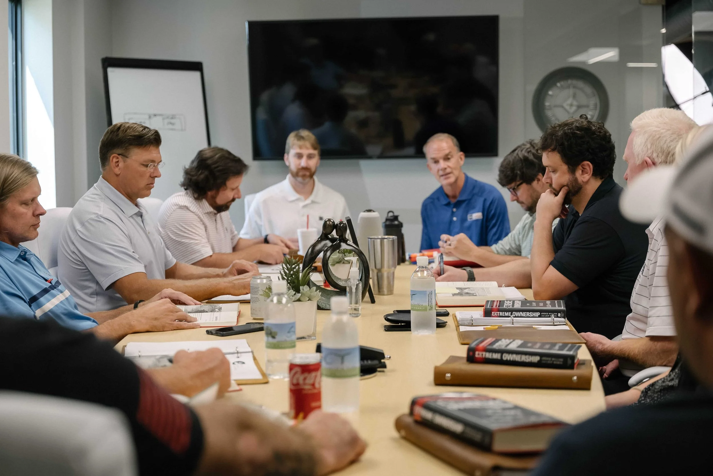 A group of people sitting around a conference table engaged in a meeting, with books, notebooks, water bottles, and beverages on the table, and a large TV screen and clock on the wall in the background.