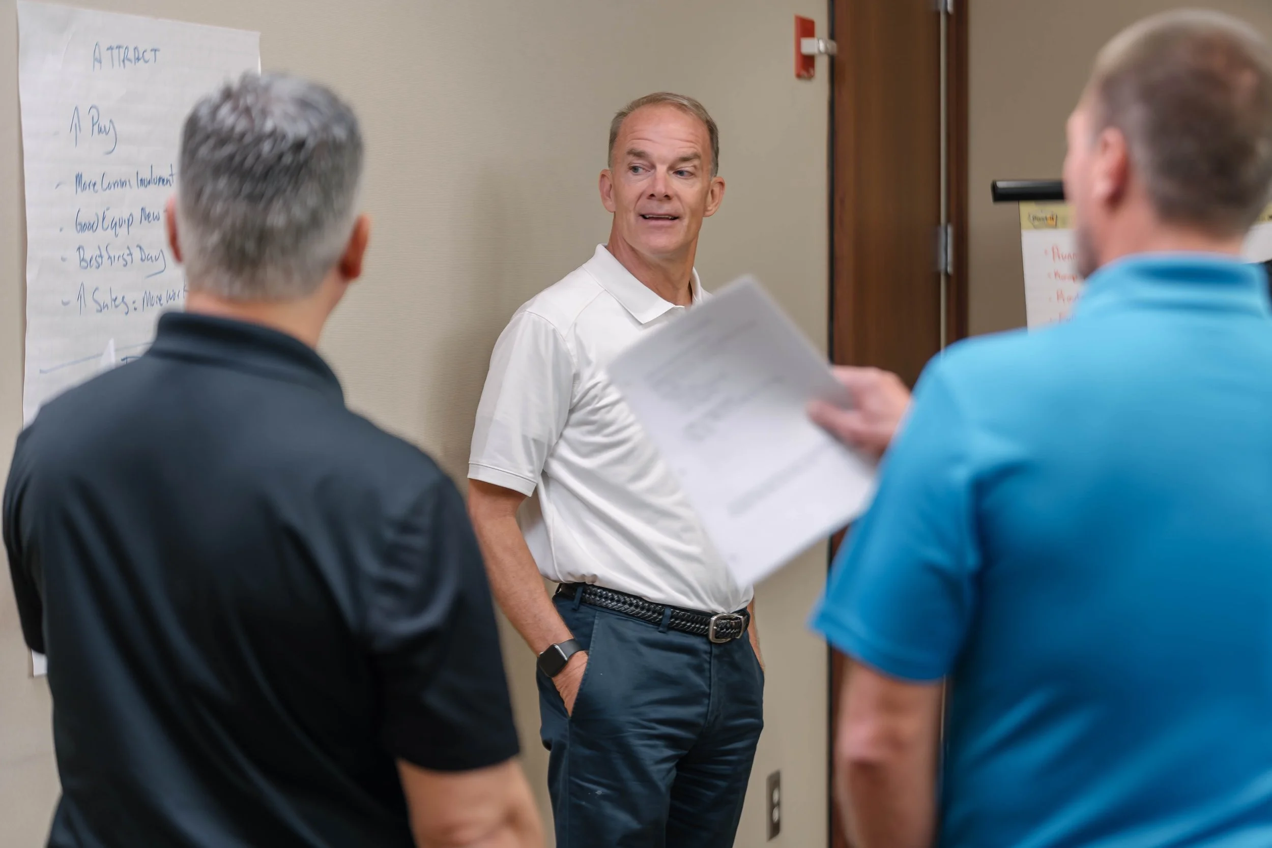 Three men in conversation in a conference room, with a man holding papers in the foreground and a whiteboard with handwritten notes in the background.