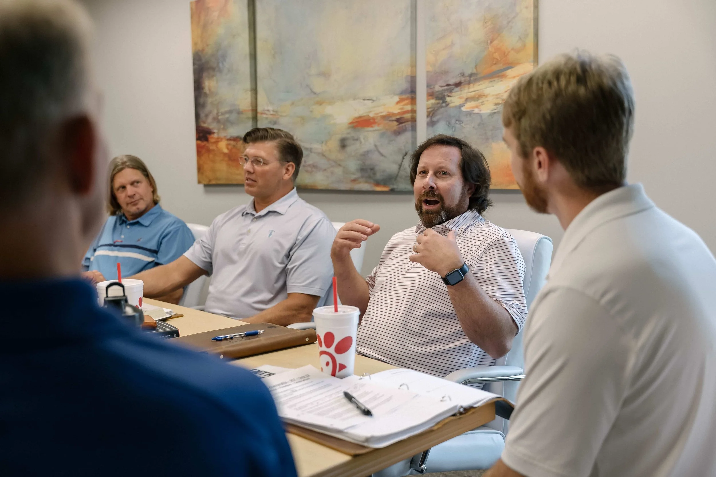 A group of five men having a discussion in a modern conference room, with artwork on the wall and Chick-fil-A drinks on the table.