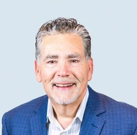 A smiling middle-aged man with gray hair and a beard, wearing a blue suit jacket and a light-colored shirt, against a light background.