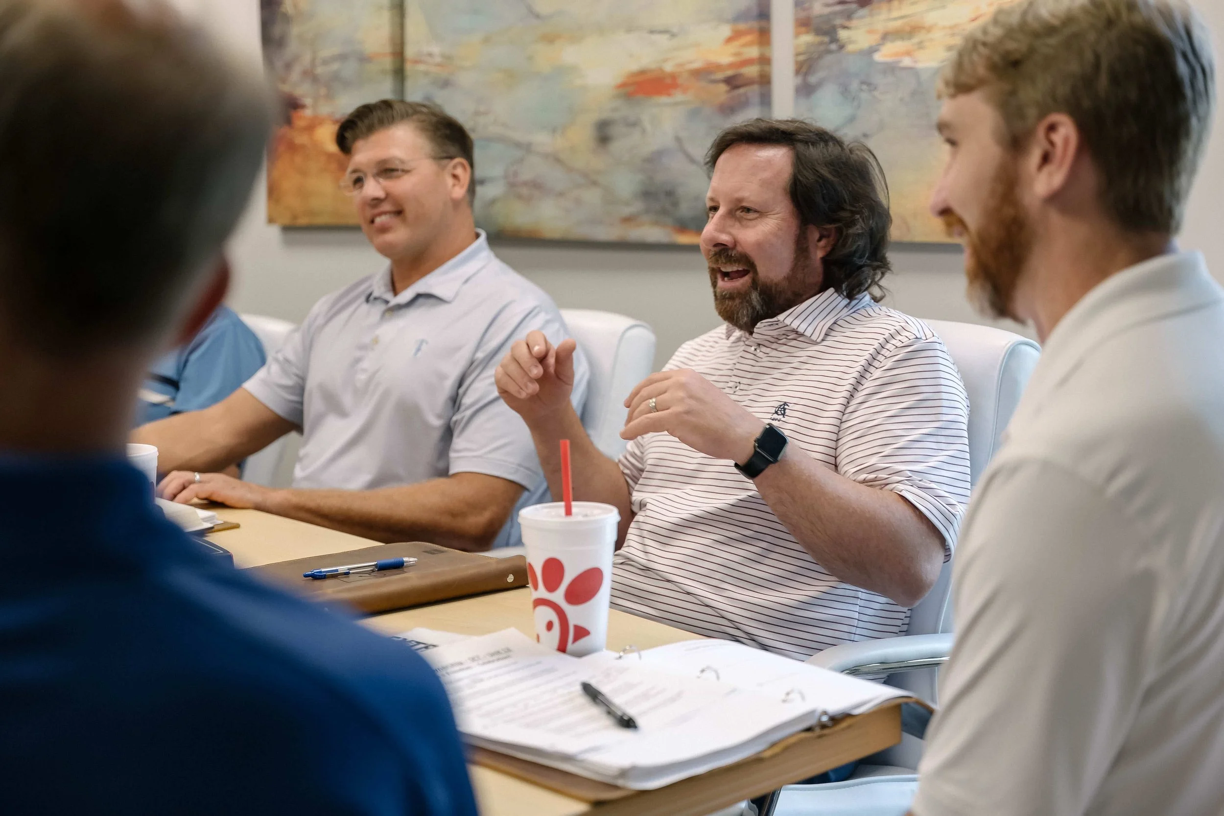 A group of men sitting at a conference table engaged in a discussion, with papers, pens, and fast food drinks in front of them.