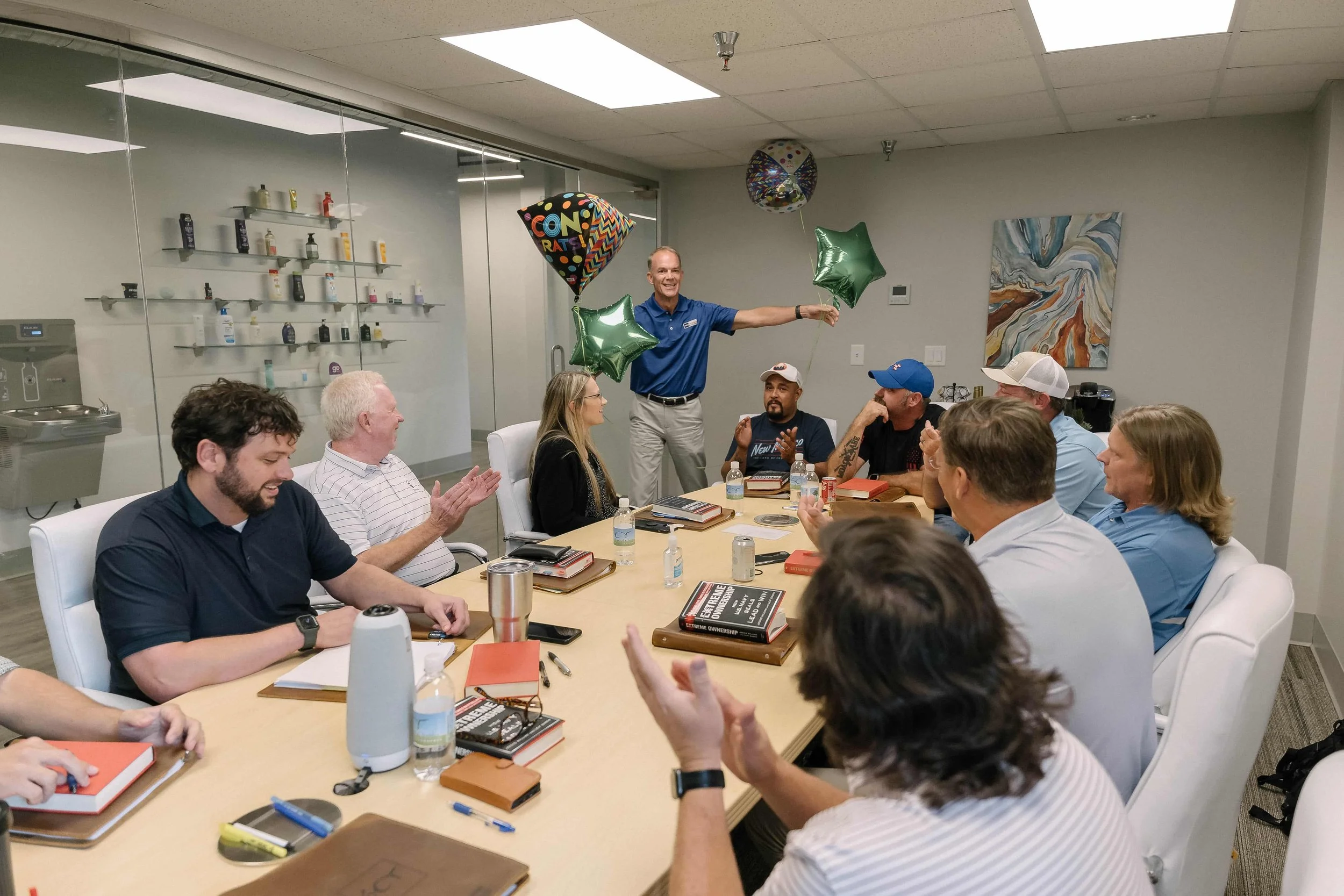A group of people sitting around a conference table, celebrating with balloons and clapping, in an office setting.