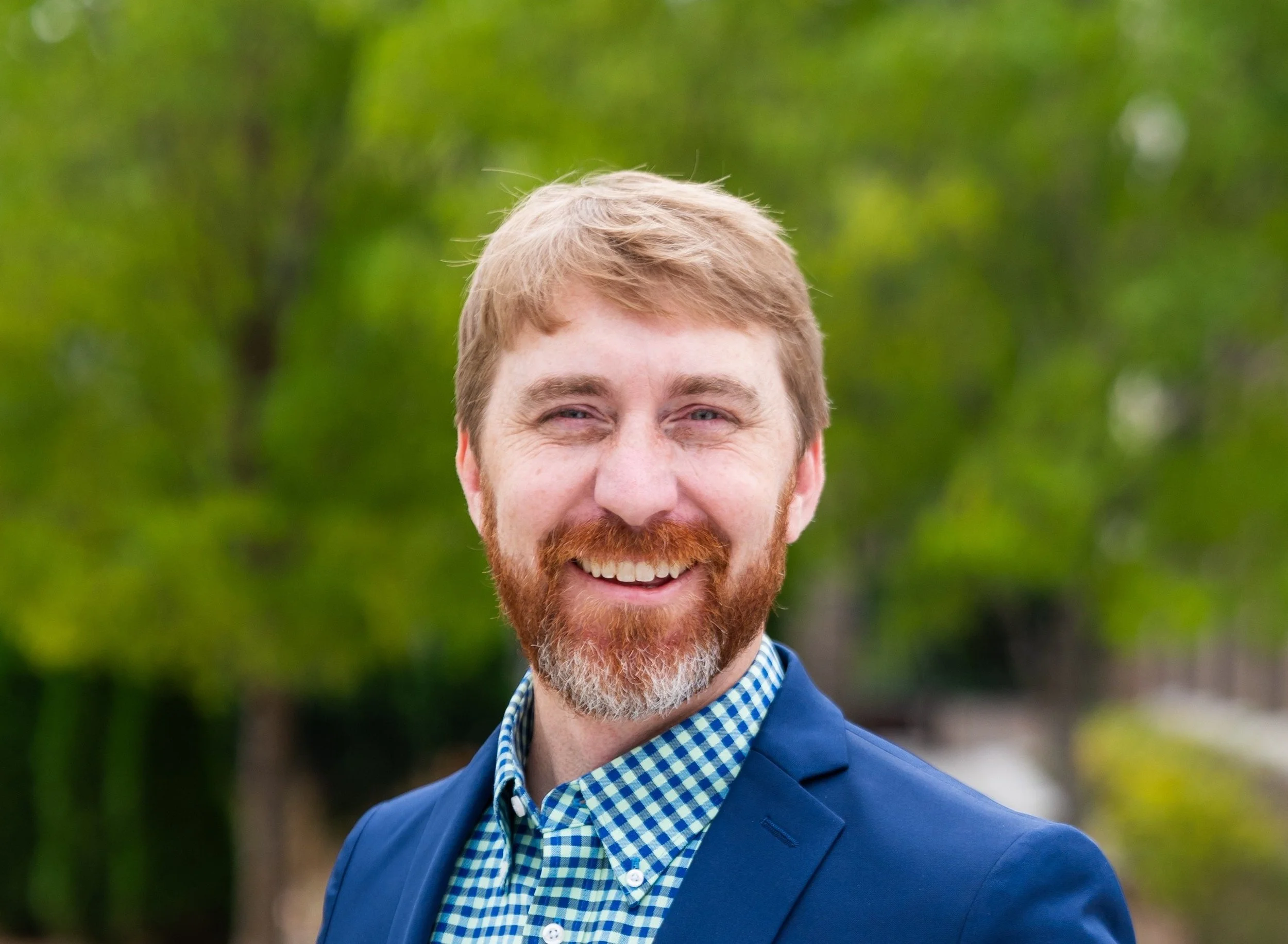 A smiling man with light brown hair, a beard, and a mustache, wearing a blue suit jacket and a blue checkered shirt, standing outdoors with blurred green trees in the background.