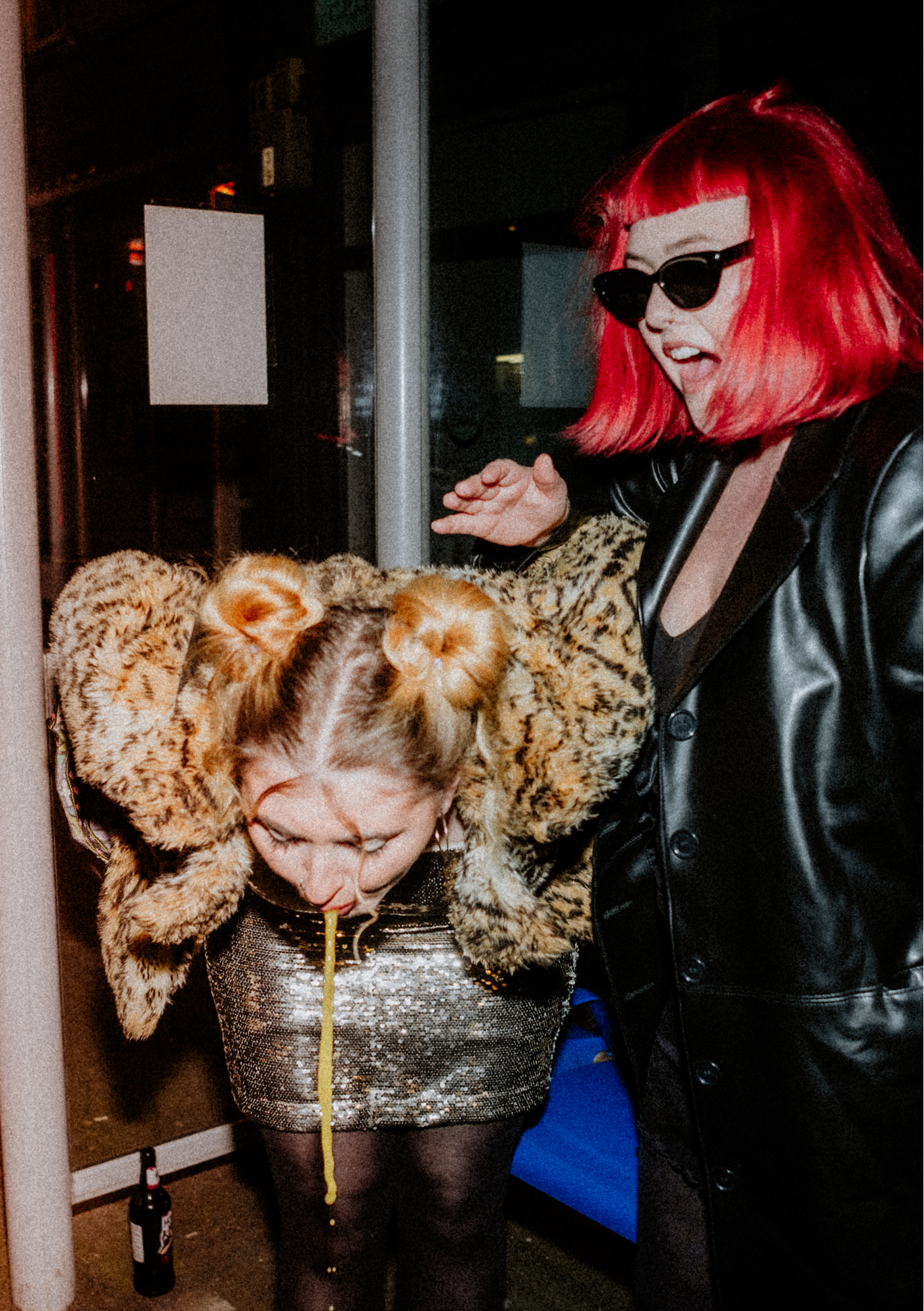 Two women celebrating a birthday. One woman wears a leopard print jacket, a silver dress, and has a long yellow spaghetti. The other woman has bright red hair, is wearing black sunglasses and a black leather jacket, and is smiling with her mouth open.