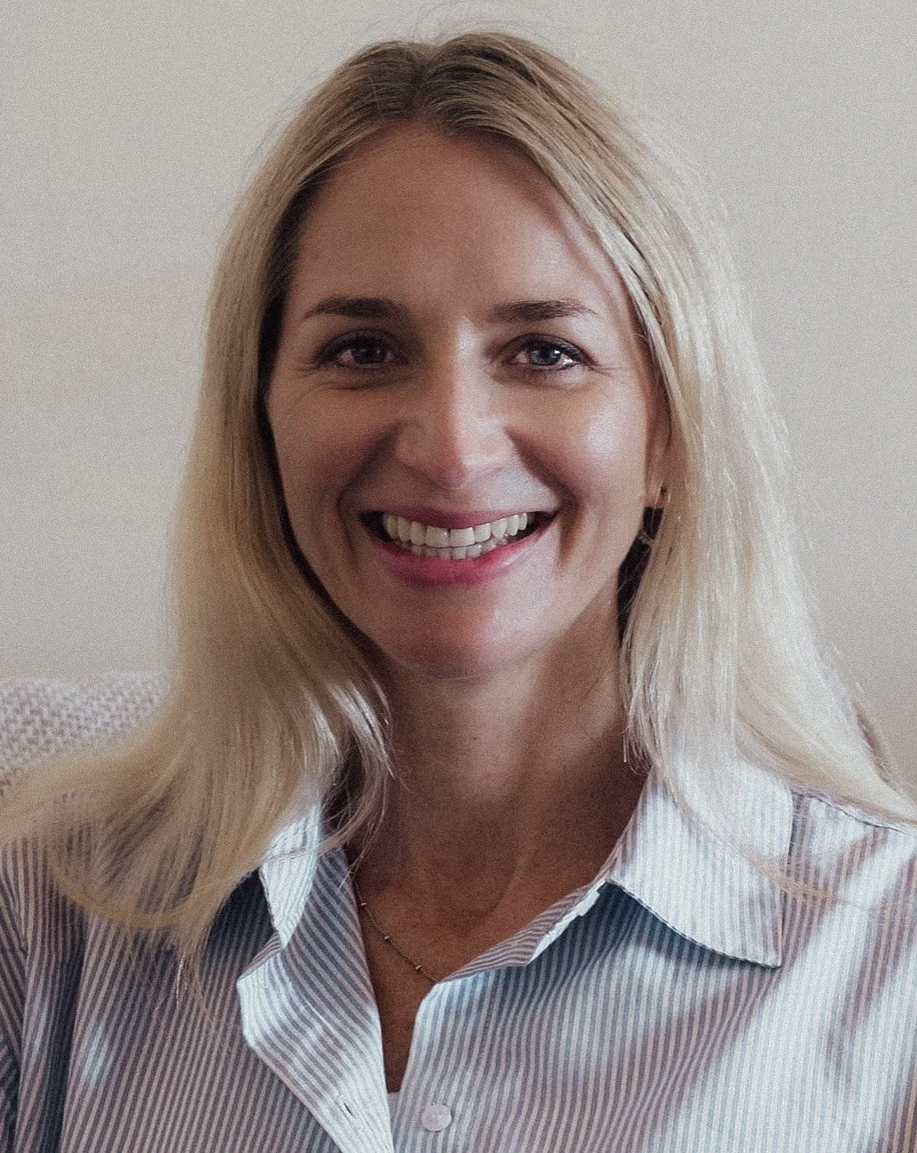 A woman with blonde hair and blue eyes smiling, wearing a striped collared shirt, against a neutral background.