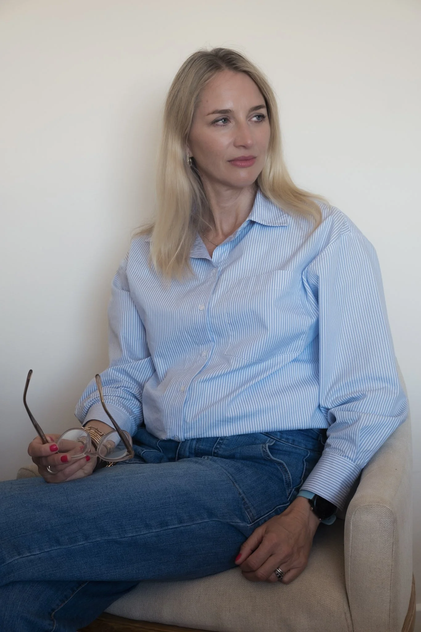 A woman with blonde hair wearing a light blue striped shirt and blue jeans sitting on a beige sofa, holding eyeglasses in her left hand and a smartwatch on her right wrist, with her hair loose and a neutral expression.