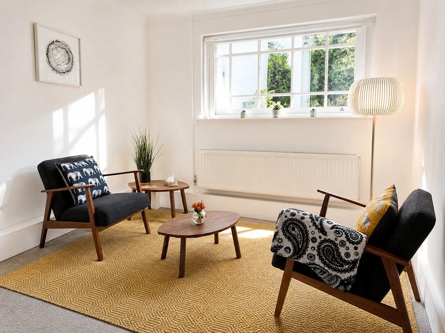 A living room with two black mid-century modern armchairs with wood arms and legs, two patterned pillows, one with an elephant design and another with yellow, a small round wooden coffee table with a vase of orange flowers, a yellow geometric patterned rug, a tall white floor lamp, a side table with a potted plant, and a large window with white trim and three small potted plants on the windowsill.
