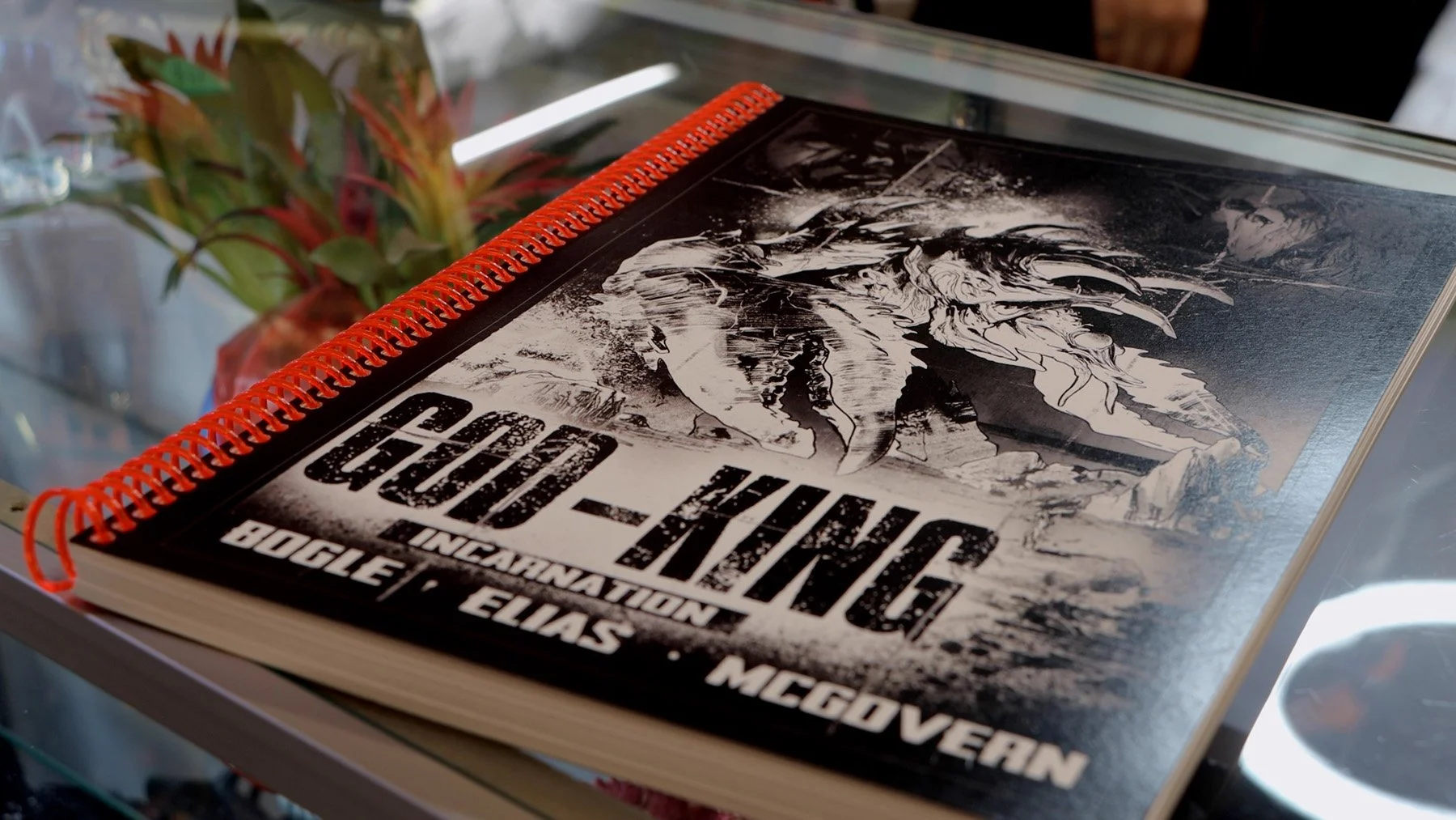 Spiral-bound preview copy of GOD-KING: Incarnation by Michael Bogle and Gabo Elias resting on a glass display case.