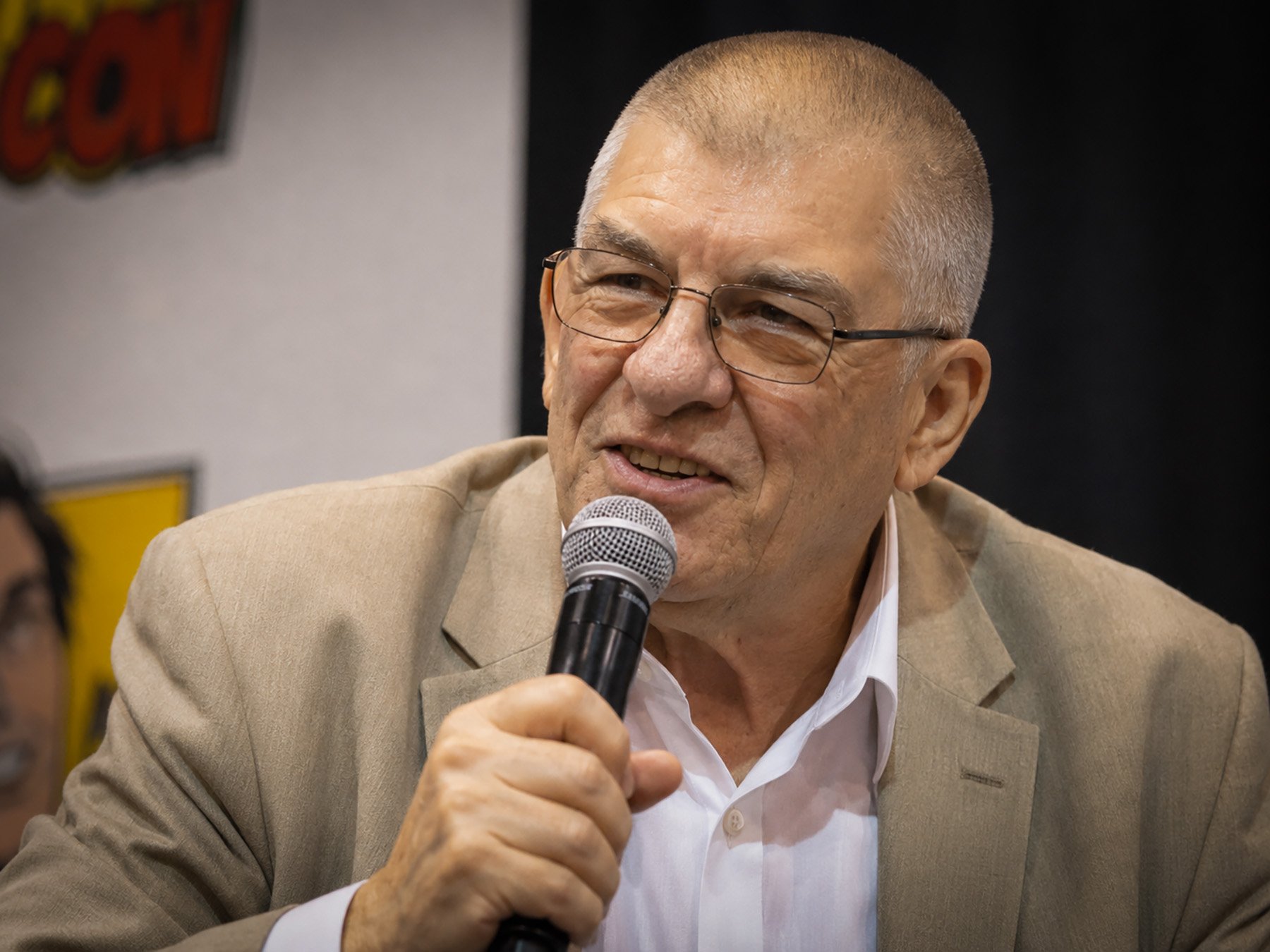 Close-up photograph of Jim Shooter speaking into a handheld microphone during a comic convention panel. He is wearing a tan blazer, white shirt and glasses while smiling as he addresses the audience.