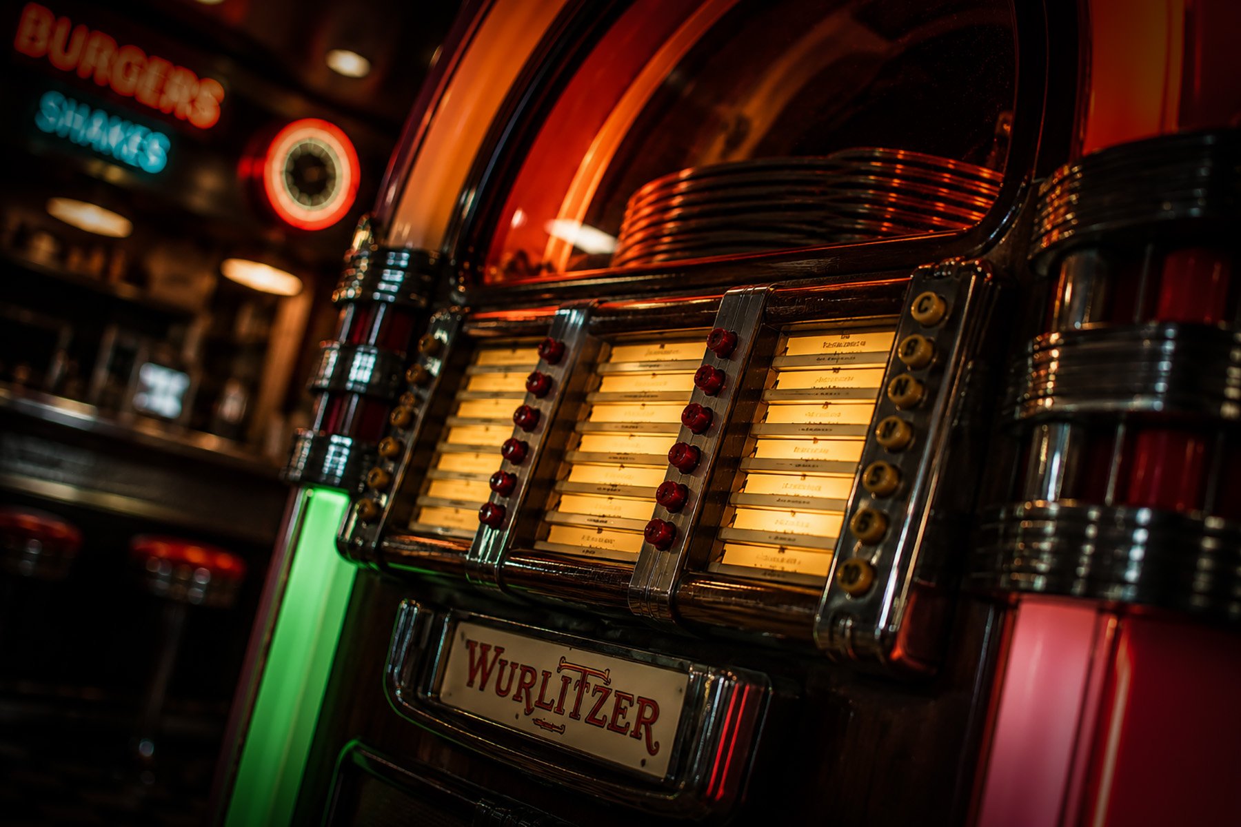 Close-up of a vintage Wurlitzer jukebox illuminated by warm neon lights inside a classic American diner, with song selection buttons and record title panels in focus.