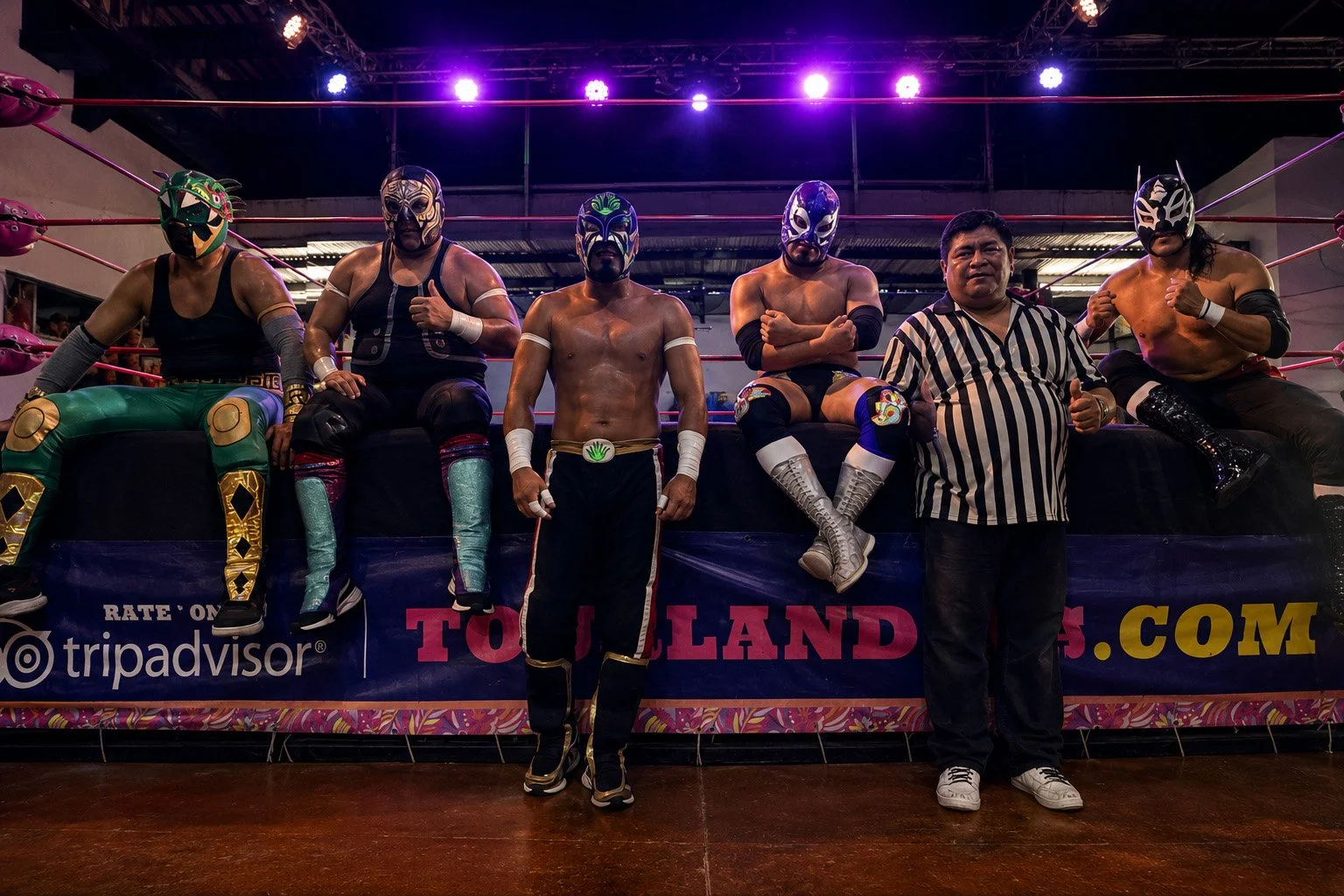 Five luchadores in colorful wrestling gear pose with a referee along the edge of a ring beneath purple arena lights.