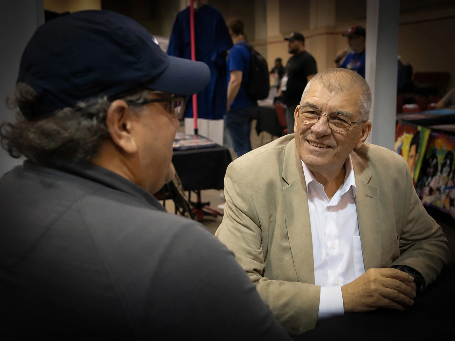 Tim San Fillippo sits across the table from Jim Shooter during Showcase Comic Con in Slidell, Louisiana. The two are smiling and engaged in conversation at Shooter's signing table while the convention continues in the background.