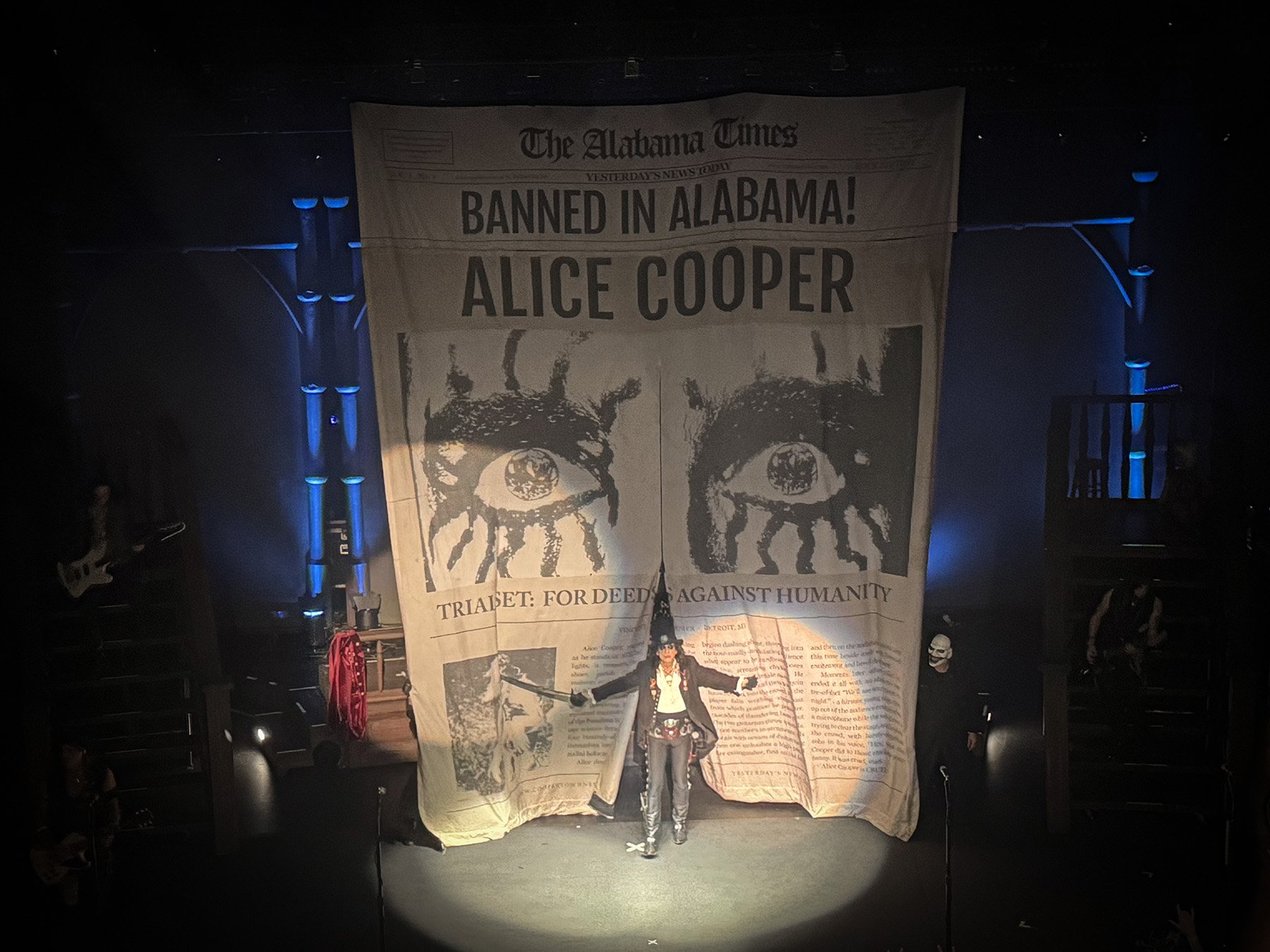 Alice Cooper stands in a spotlight onstage at the Saenger Theatre in Mobile, Alabama, beneath a giant theatrical newspaper backdrop reading “Banned in Alabama! Alice Cooper.”