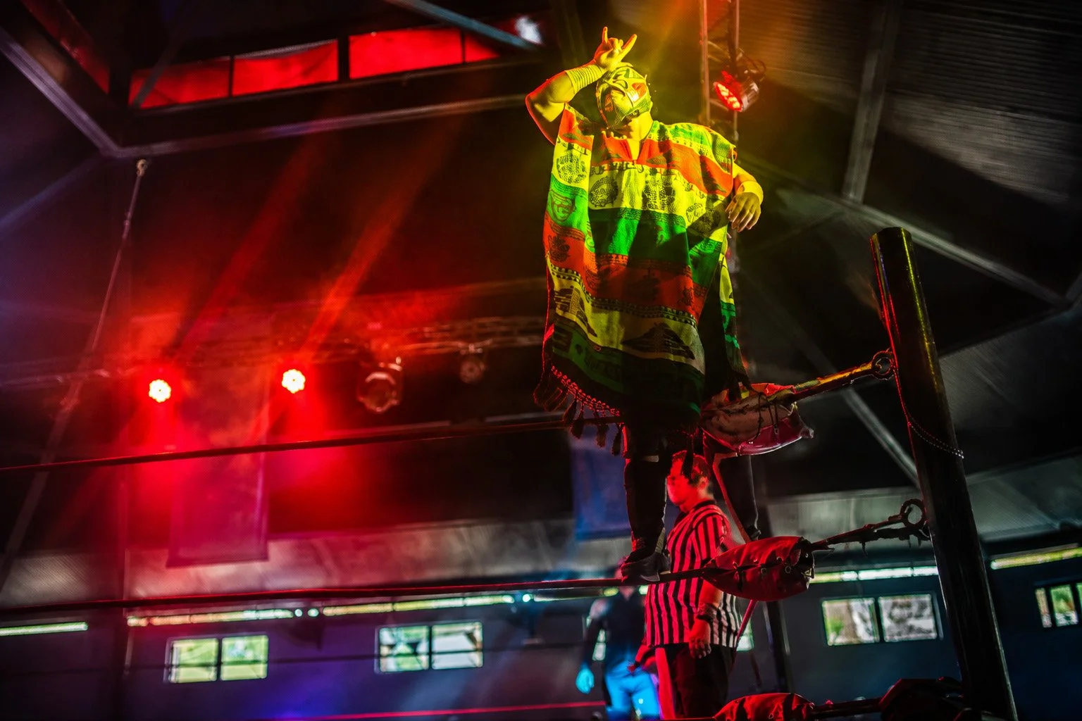 Masked luchador in a colorful poncho standing atop a ring turnbuckle under dramatic red and yellow arena lights.