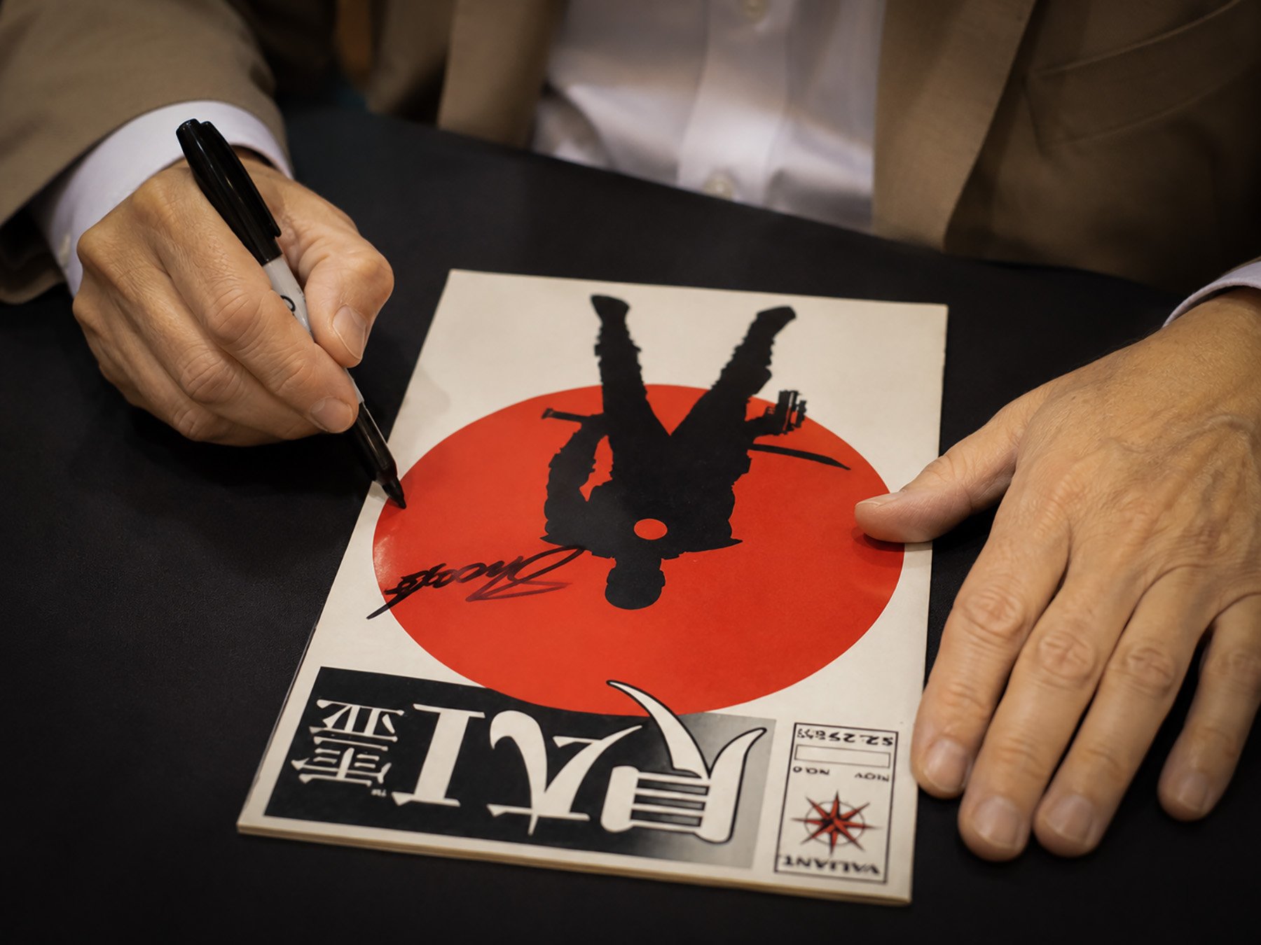 Close-up of Jim Shooter signing a copy of Rai #0 with a black marker at a convention table. The comic's bold black silhouette and red sun cover fill the frame as his hands rest on either side of the book.