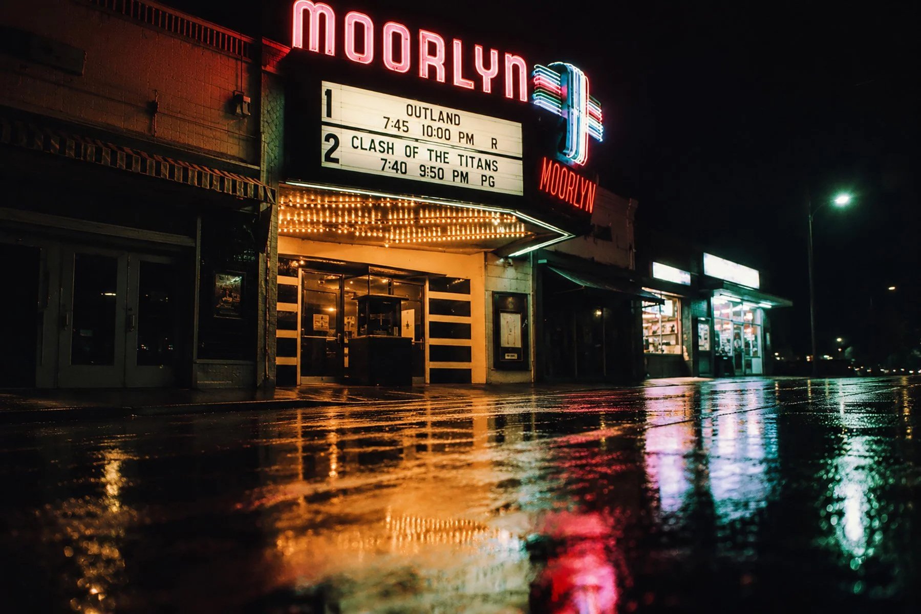 The historic Moorlyn Theatre illuminated at night, with neon lights reflecting across a rain-soaked street beneath the classic movie marquee.