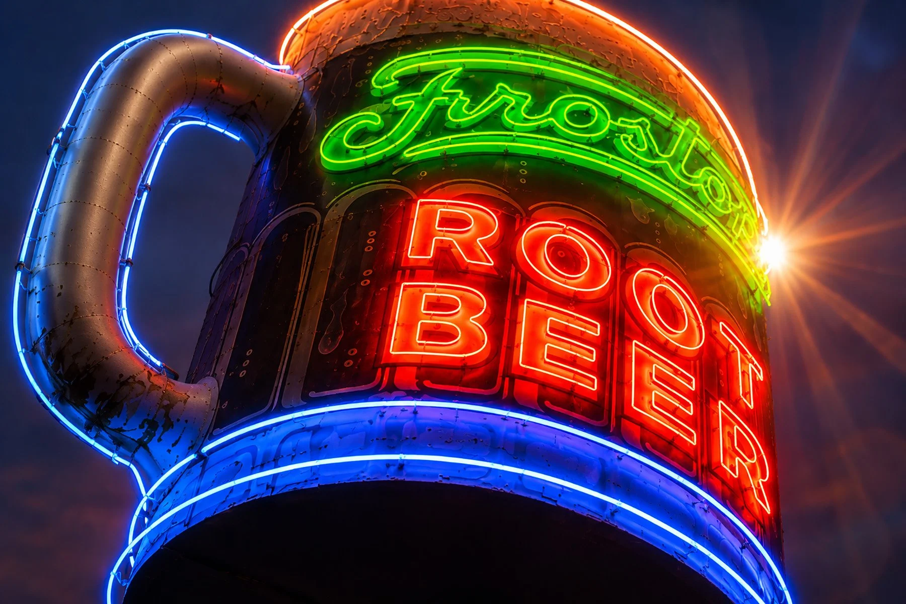 Close-up of the illuminated vintage Frostop Root Beer mug sign outside Ted's Frostop in New Orleans at night, featuring bright green, red and blue neon against the evening sky.