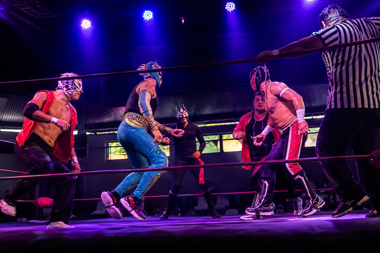 Five luchadores crowd the center of the ring while a referee monitors the confrontation beneath purple arena lights.
