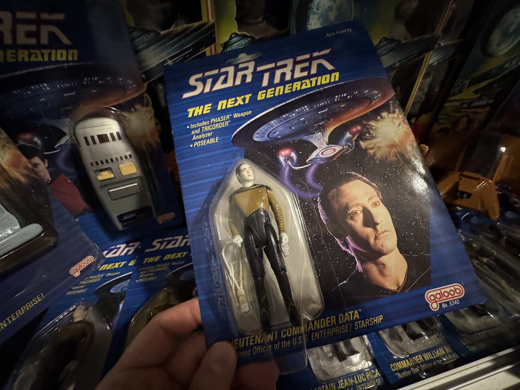 Carded Galoob Lieutenant Commander Data "Blue Face" variant from Star Trek: The Next Generation held in front of a display featuring other carded Galoob figures, the phaser, communicator and vehicles.