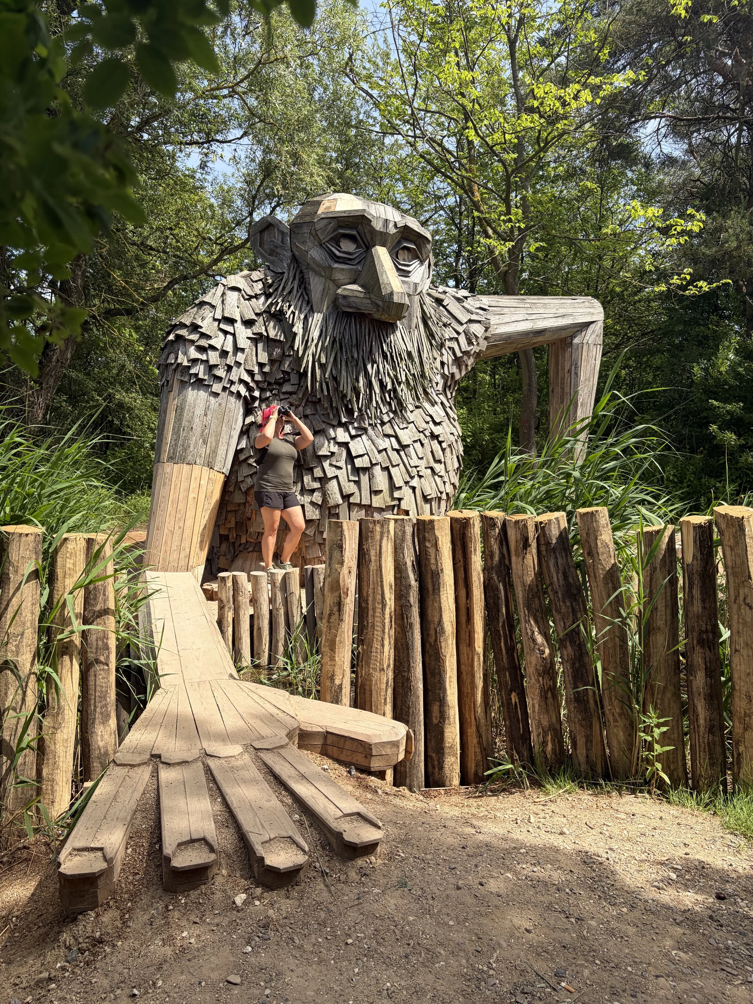 A large wooden sculpture of a troll is situated in a forested area, with a wooden walkway leading towards it. A woman is standing in front of the sculpture, looking with binoculars, surrounded by lush green trees and plants.